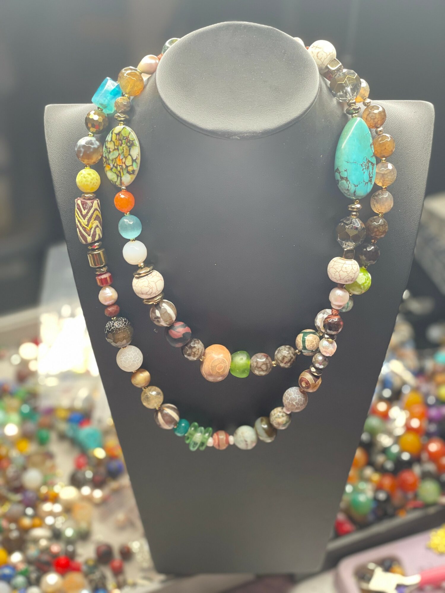 Two layered beaded necklaces with colorful and patterned beads displayed on a black bust, background filled with more jewelry pieces.