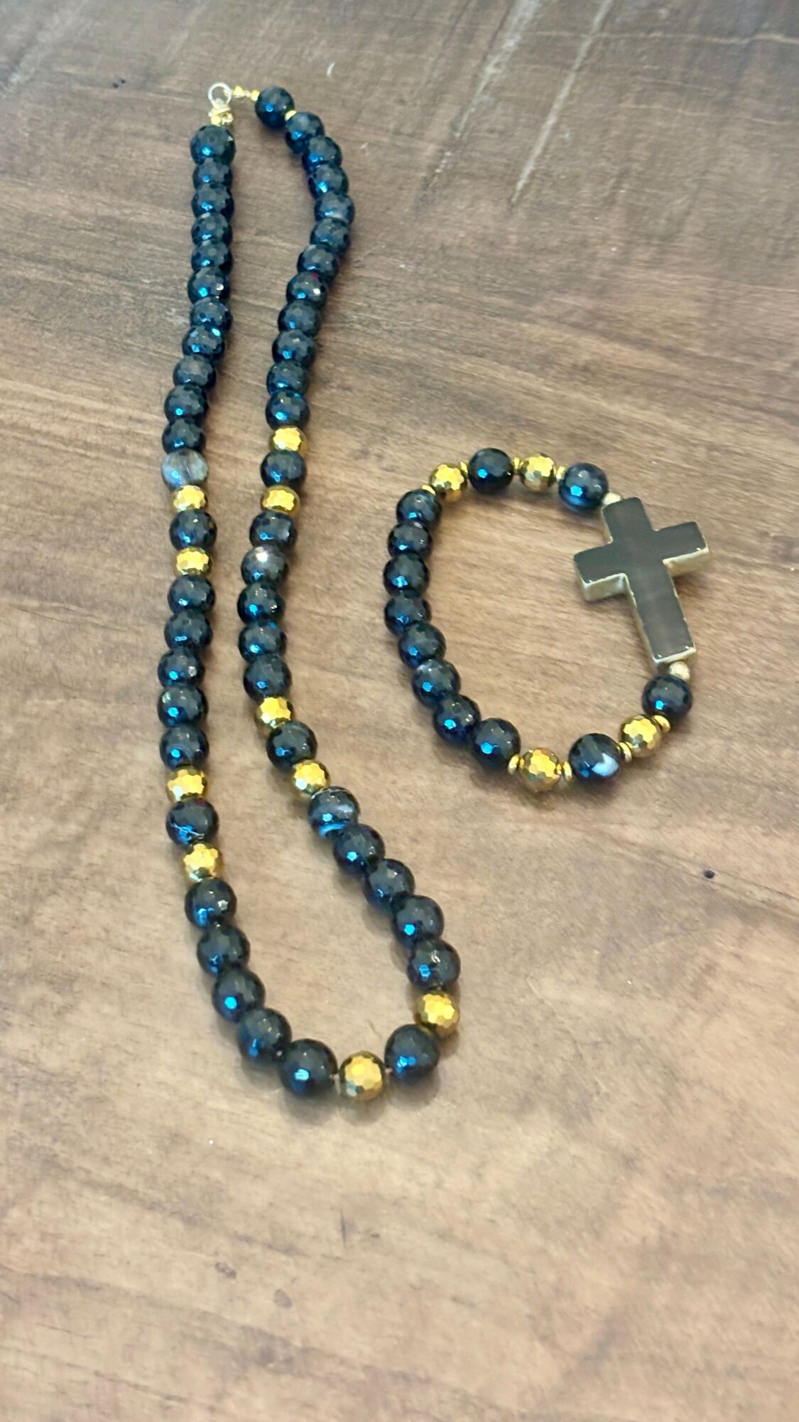 A beaded necklace and bracelet with blue and gold beads, the bracelet has a cross pendant, on a wooden surface.