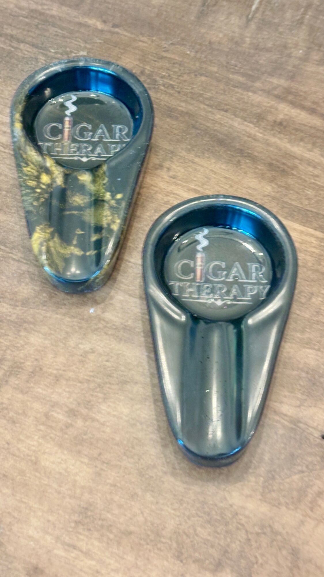 Two cigar cutters on a wooden surface, one with a blue top and gold design, the other silver, both with clear plastic covers.