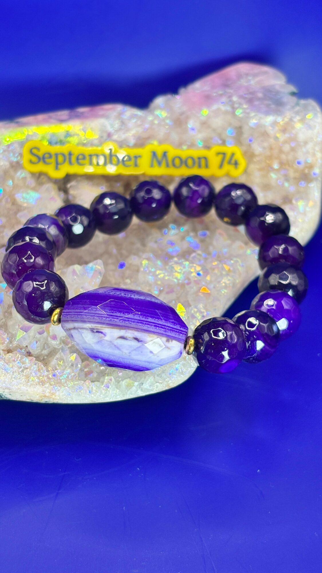 Purple beaded bracelet with a large central stone, resting on a glittery surface with a blue background.