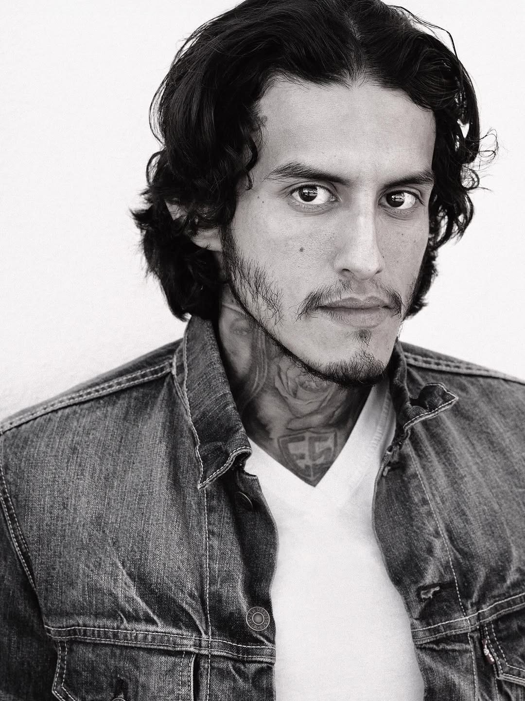 Portrait of a man with wavy dark hair, wearing a denim jacket and white shirt, looking at the camera.