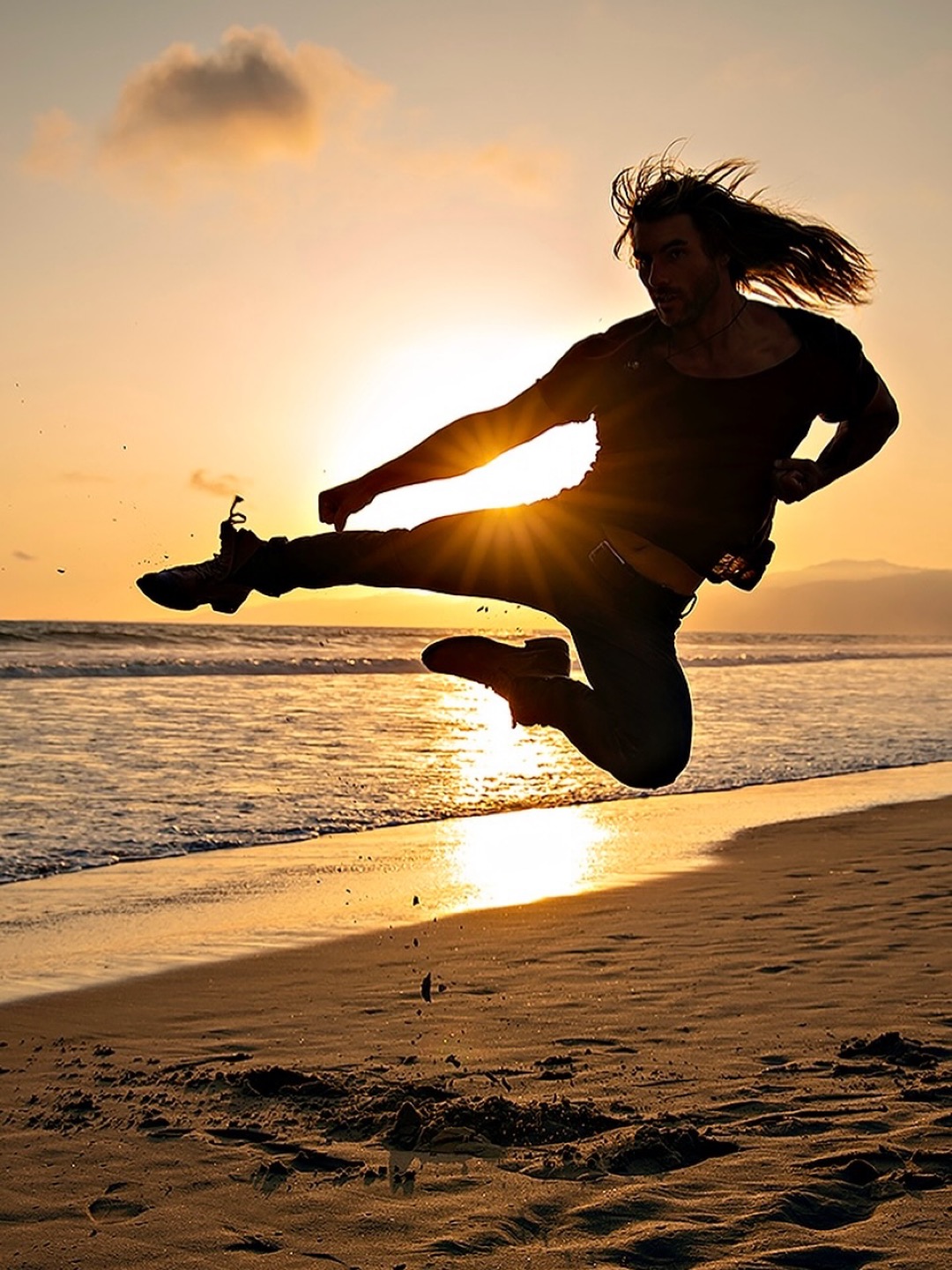 Person jumping on beach during sunset with hair flowing, silhouette against the sun, ocean and sky in background.