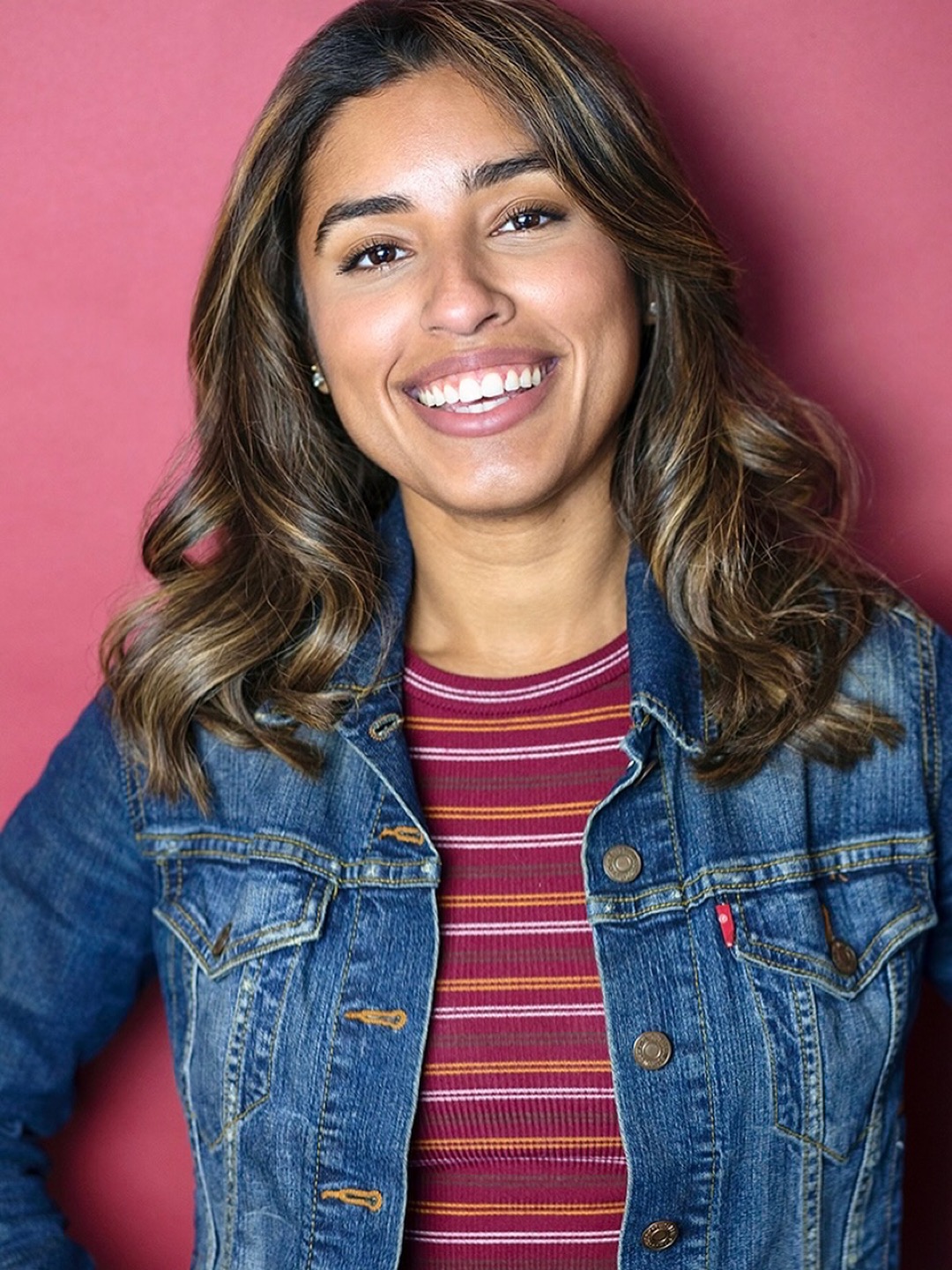 Young woman with wavy hair smiling, wearing a denim jacket and striped shirt, against a pink background.