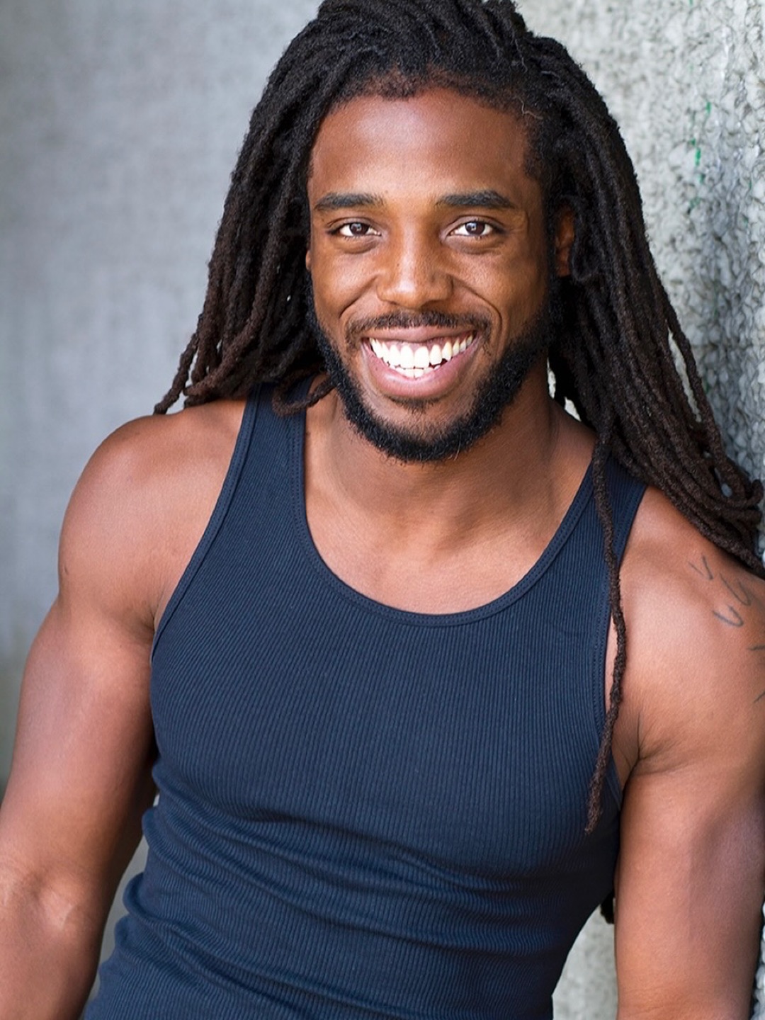 Smiling man with long dreadlocks wearing a navy tank top, standing outdoors against a gray background.