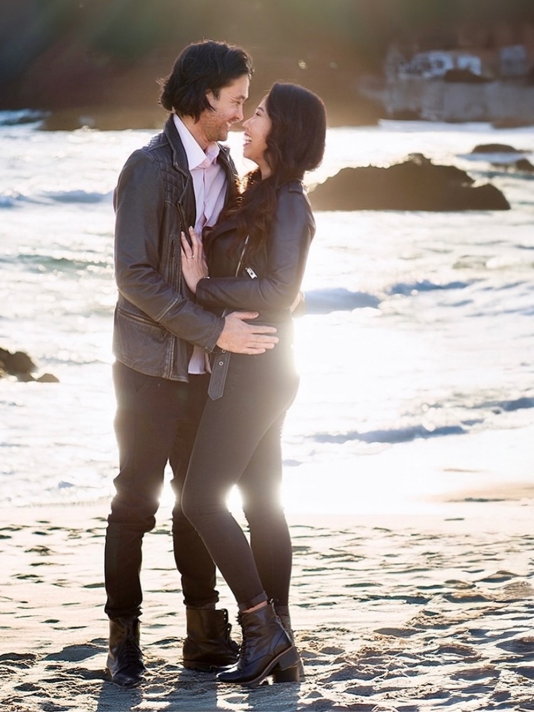 A couple stands close together on a beach, facing each other with the ocean and rocks in the background.