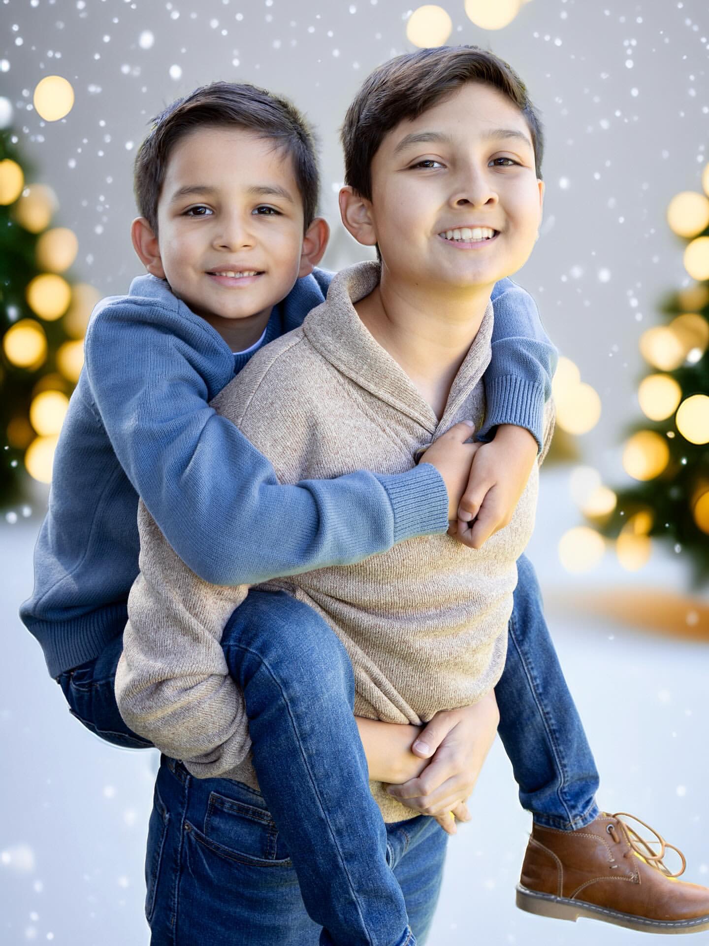 Two boys smiling, one hugging the other from behind, in front of a festive background with snow and lights.