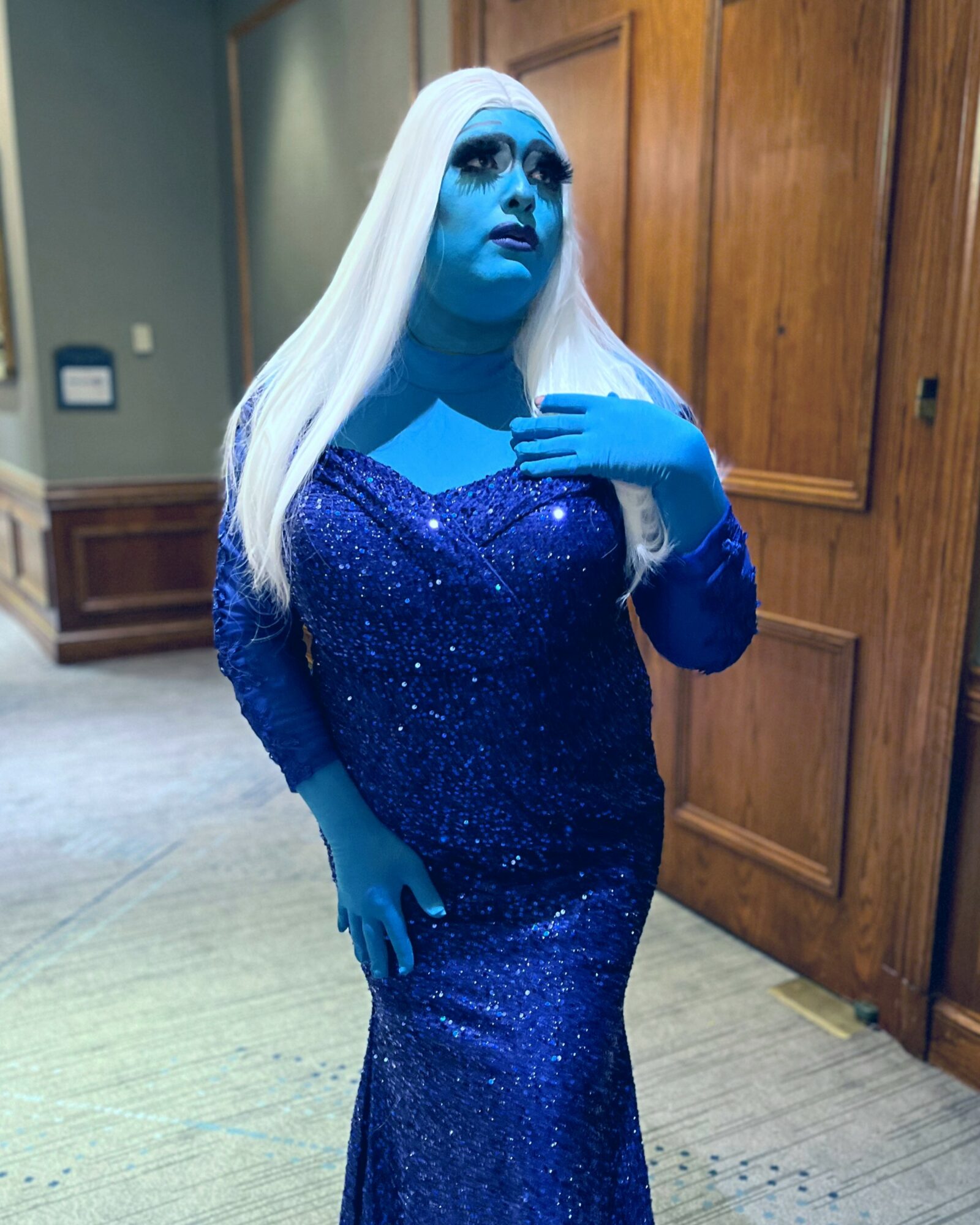 Person with blue skin and long white hair wearing a sparkly blue dress, standing indoors near wooden panels.
