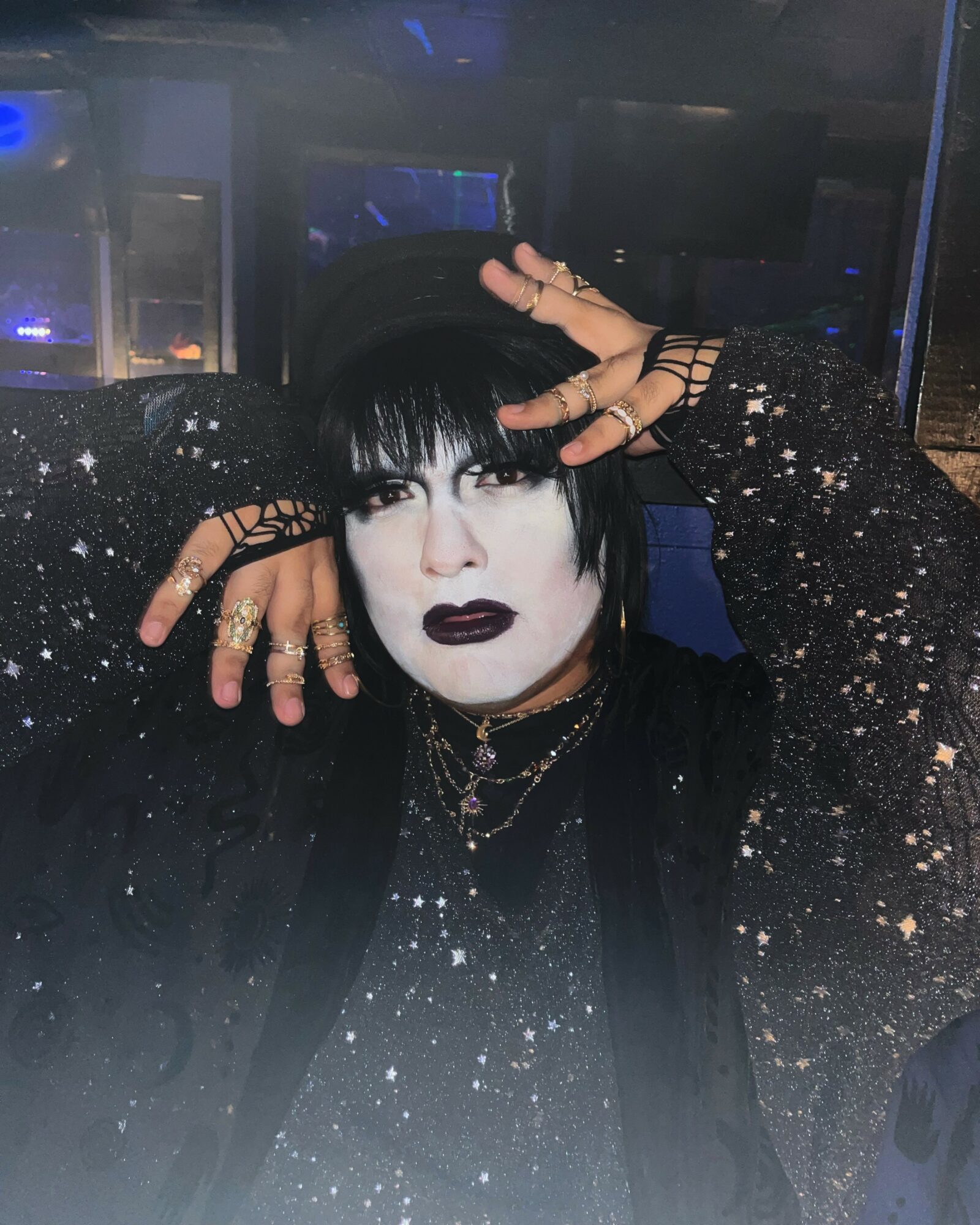 Person with dark makeup, black hair, and glittery clothing poses with hands near face in a dark setting.