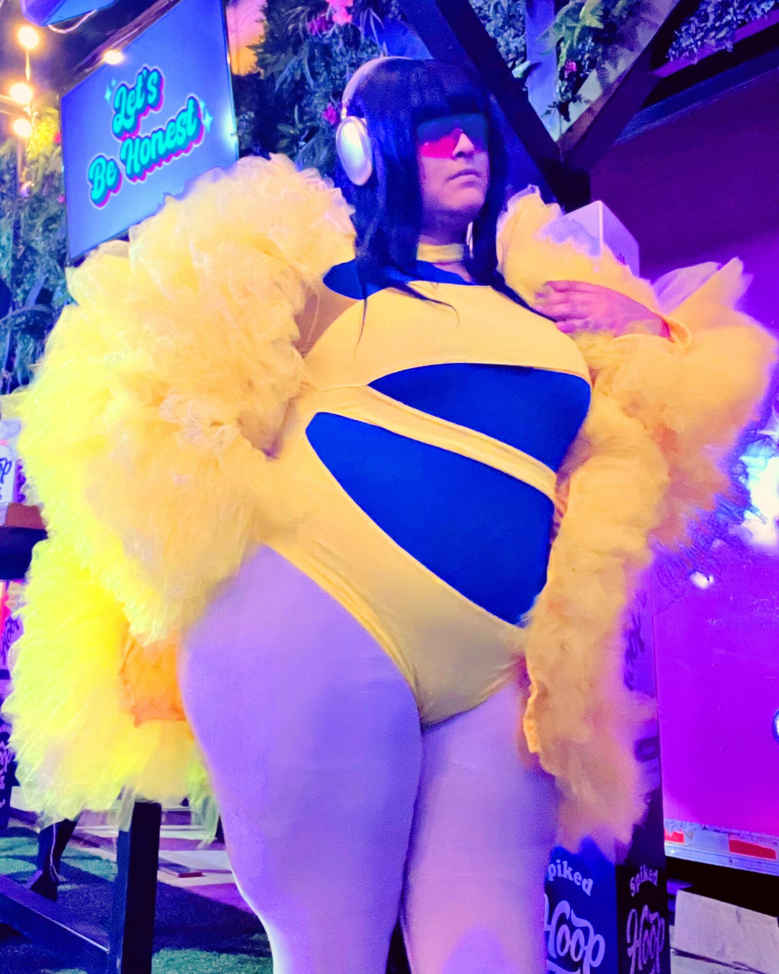 Person in yellow and black costume with large yellow fluffy sleeves, wearing headphones and sunglasses, standing outdoors at night.