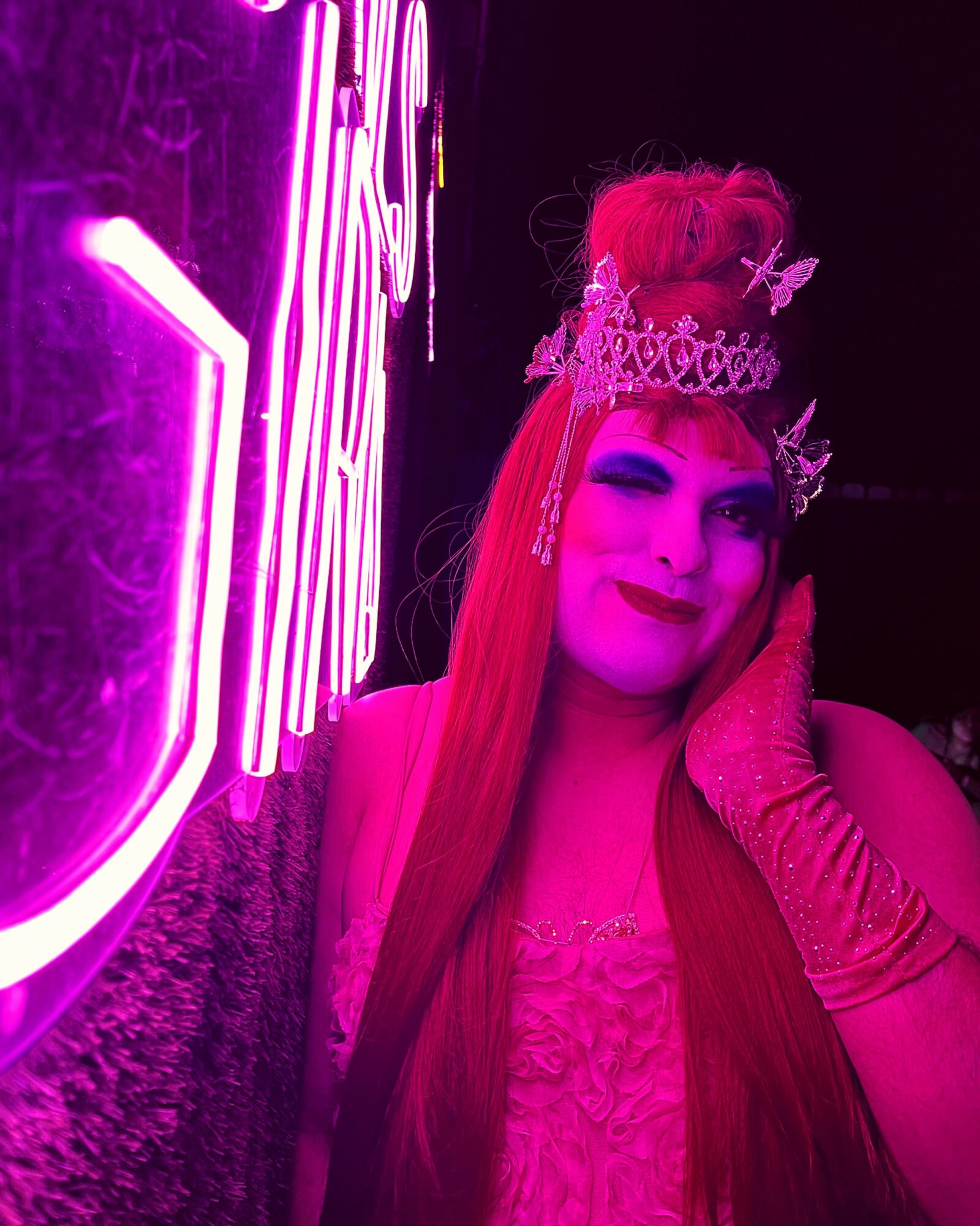 Person with long hair and crown, smiling, illuminated by pink neon light, touching face with gloved hand.