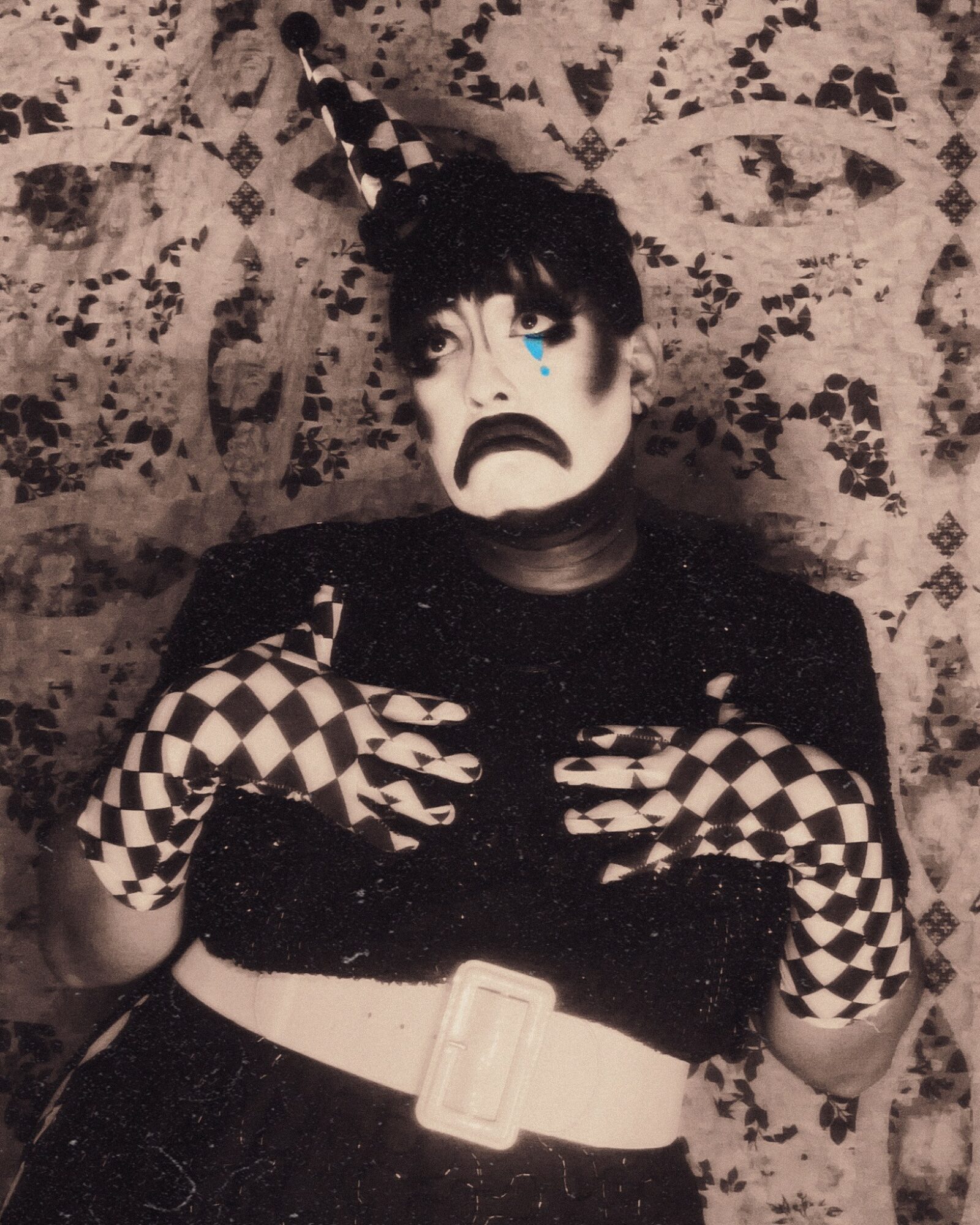 Person with clown makeup, checkered gloves, and a hat, sitting against a patterned background, with a sad expression.