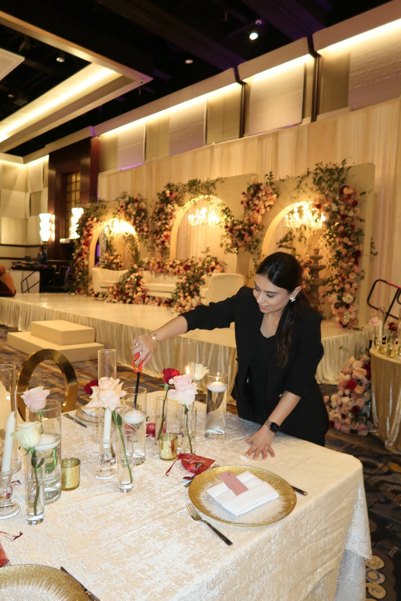 Person setting a table with glasses and decorations in a decorated event space with floral arrangements and warm lighting.