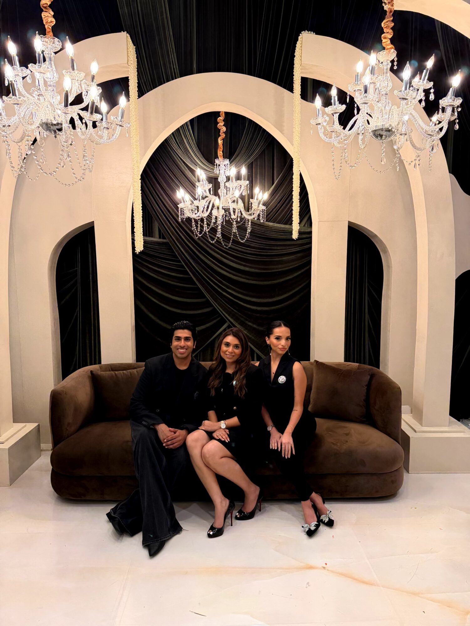 Three people sitting on a brown sofa in front of a black and white backdrop with chandeliers and drapery, in an elegant setting.