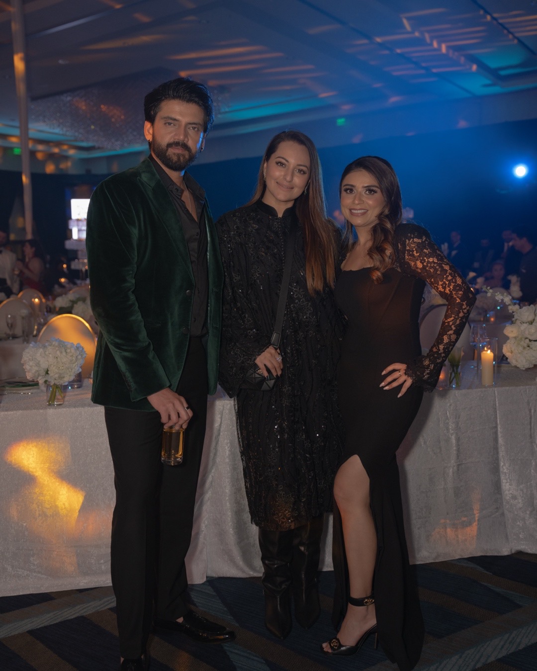 Three people dressed in formal attire at a party, standing in front of a decorated table with candles and flowers.