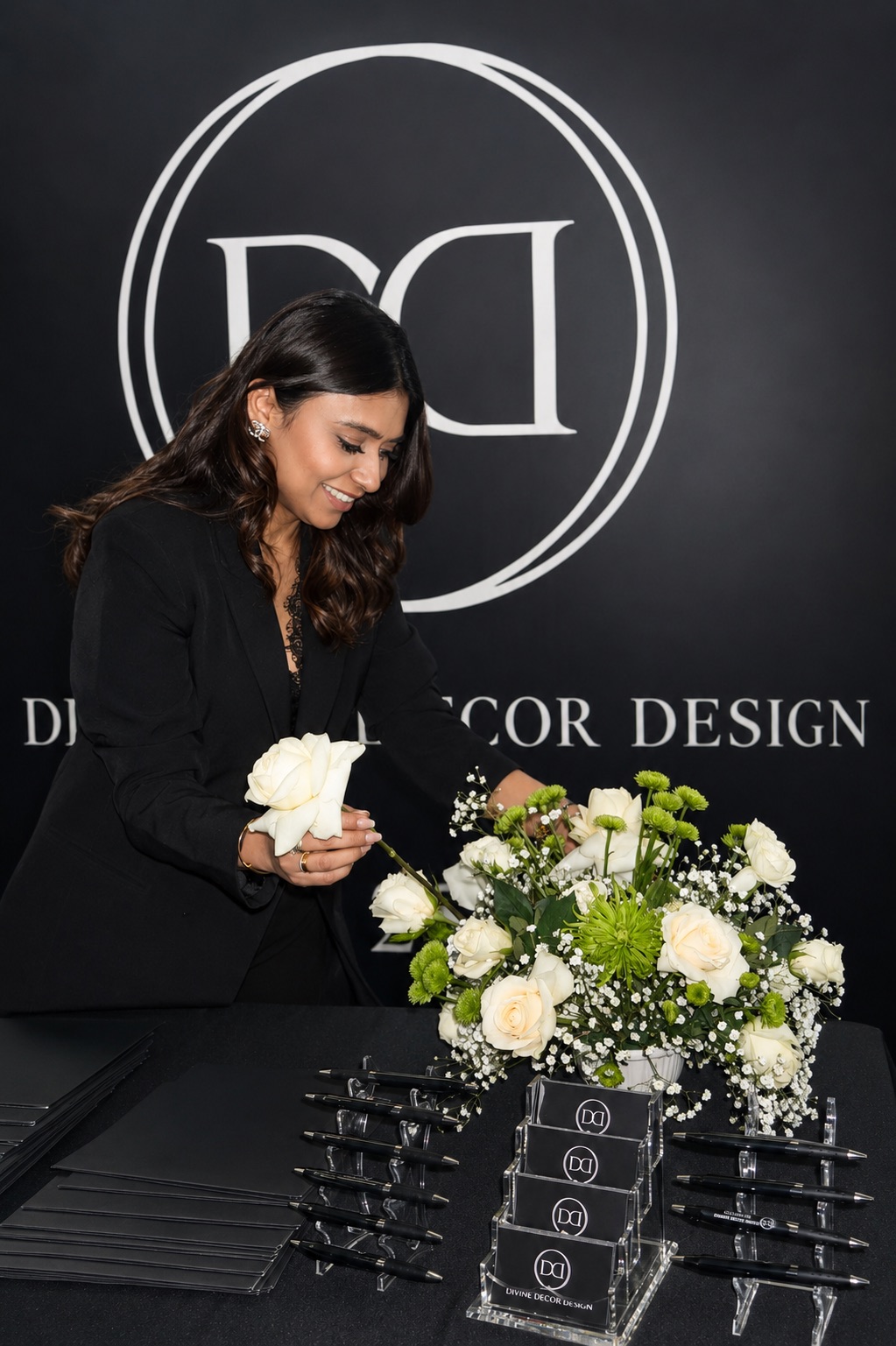 Woman arranging white and green flowers on table in front of a black backdrop with logo and text.