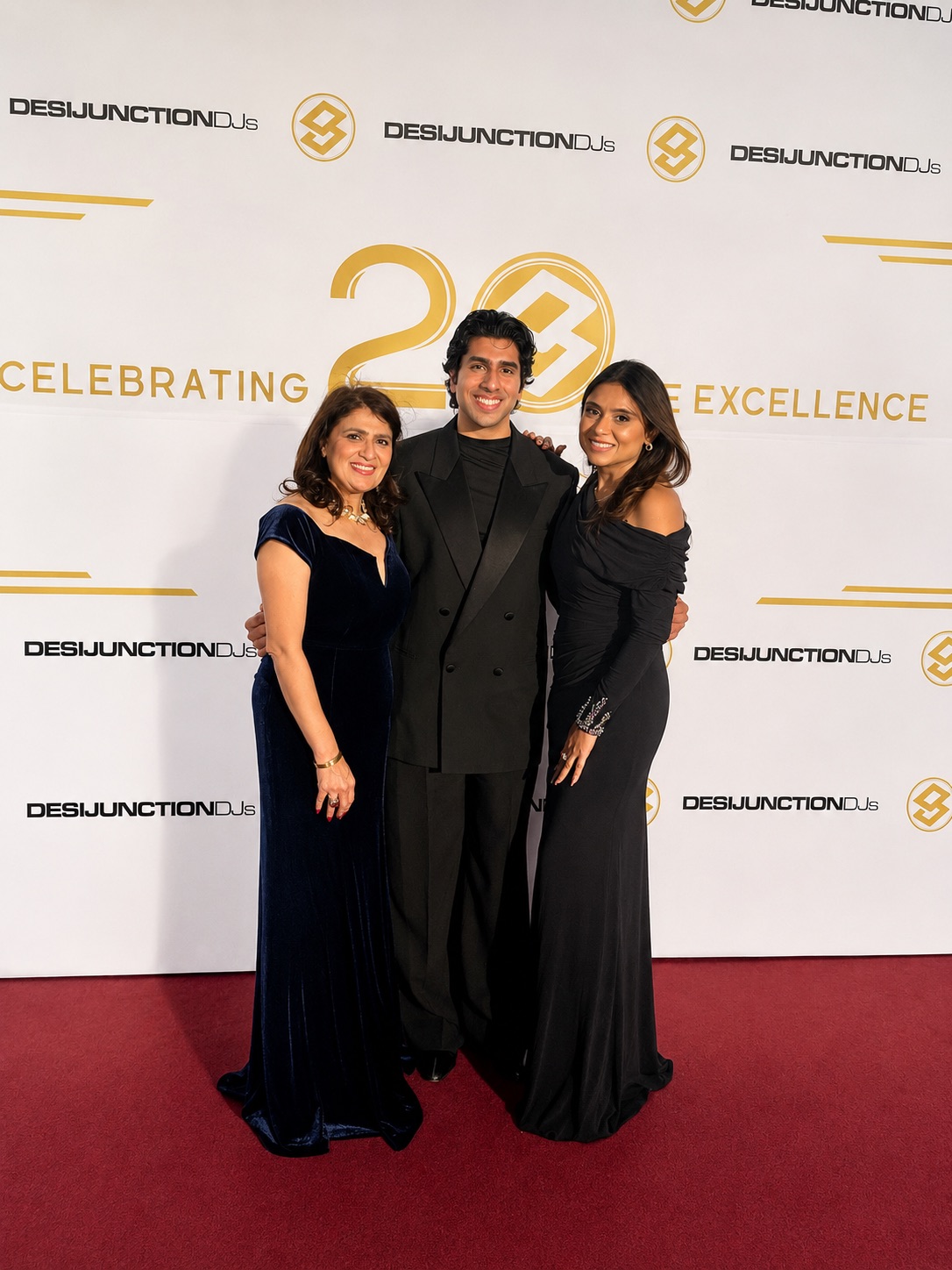 Three people in formal attire standing on a red carpet in front of a backdrop with logos and text, smiling.