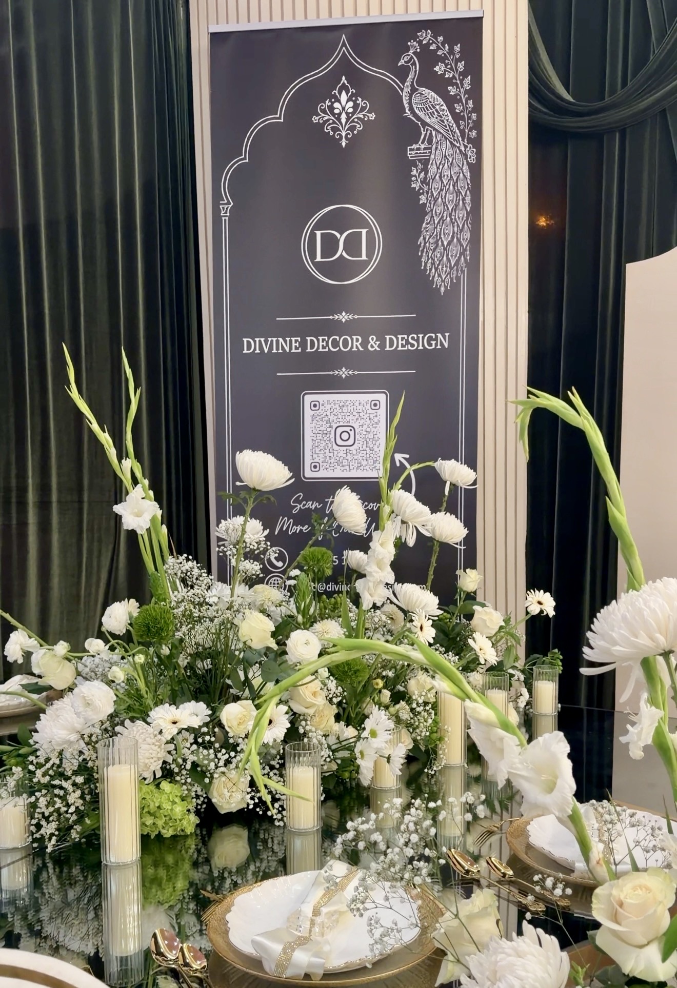 Decorative floral arrangement with white flowers and candles in front of a sign for Divine Decor & Design, featuring a QR code.