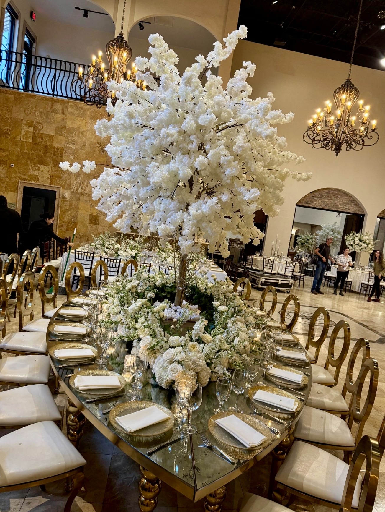 Round table with a large white floral centerpiece and place settings, in an elegant banquet hall with chandeliers.