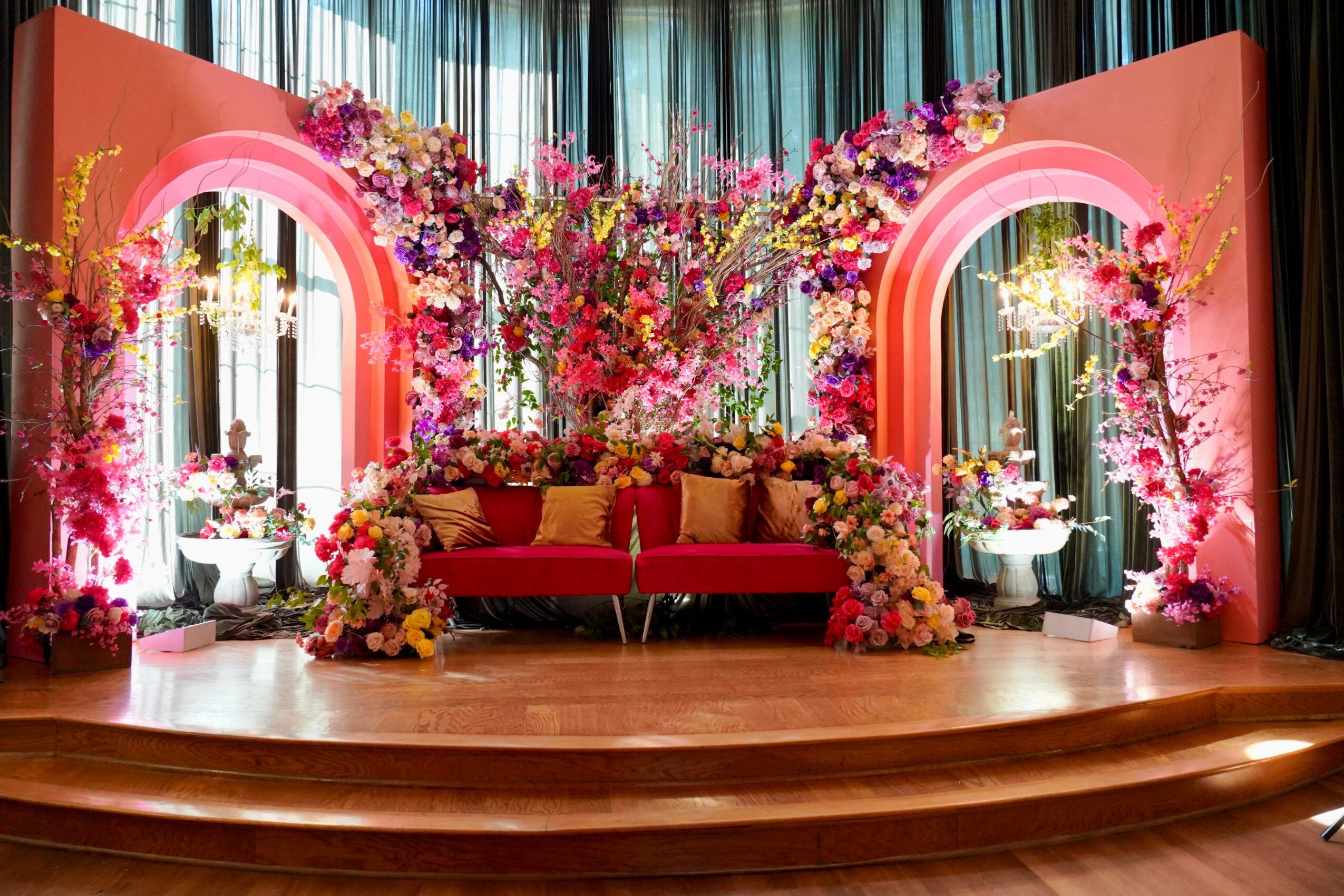 Stage decorated with pink floral arrangements, a red sofa, and pink arches, with curtains in the background.