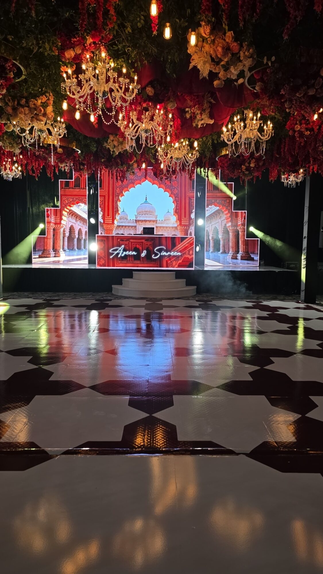 Stage decorated with flowers and chandeliers, with a backdrop of an arch and red curtains, reflecting on a shiny floor.