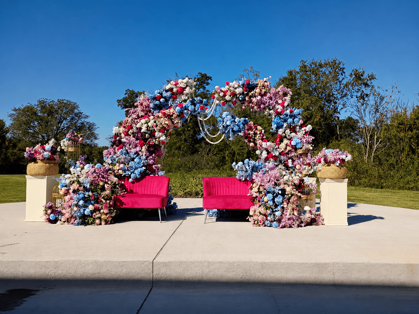 Decorative floral arch with pink, purple, and white flowers, two pink chairs, and flower arrangements on pedestals outdoors.