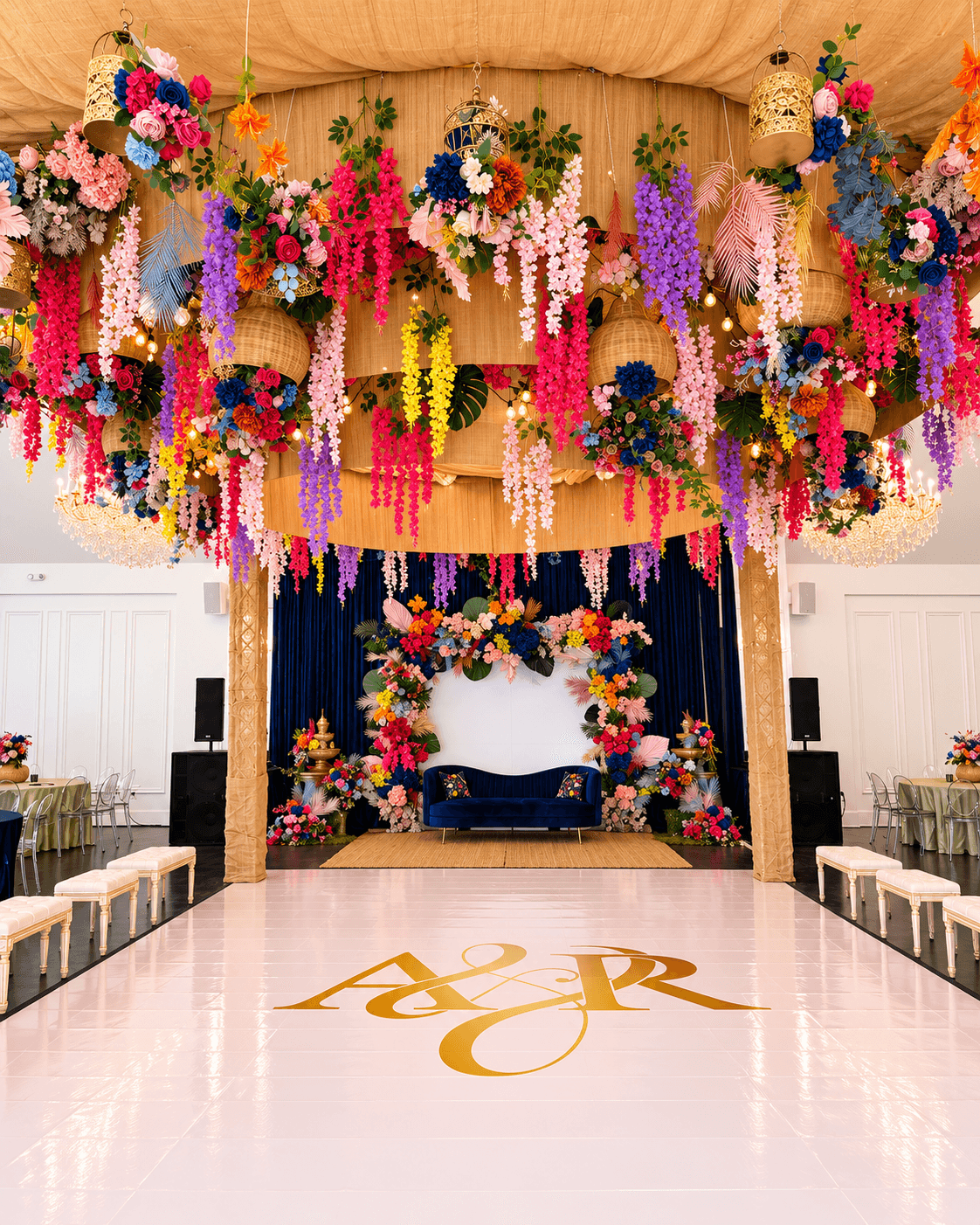 Colorful floral arrangements hang from a ceiling, with a decorated stage and a white backdrop, in a decorated event space.