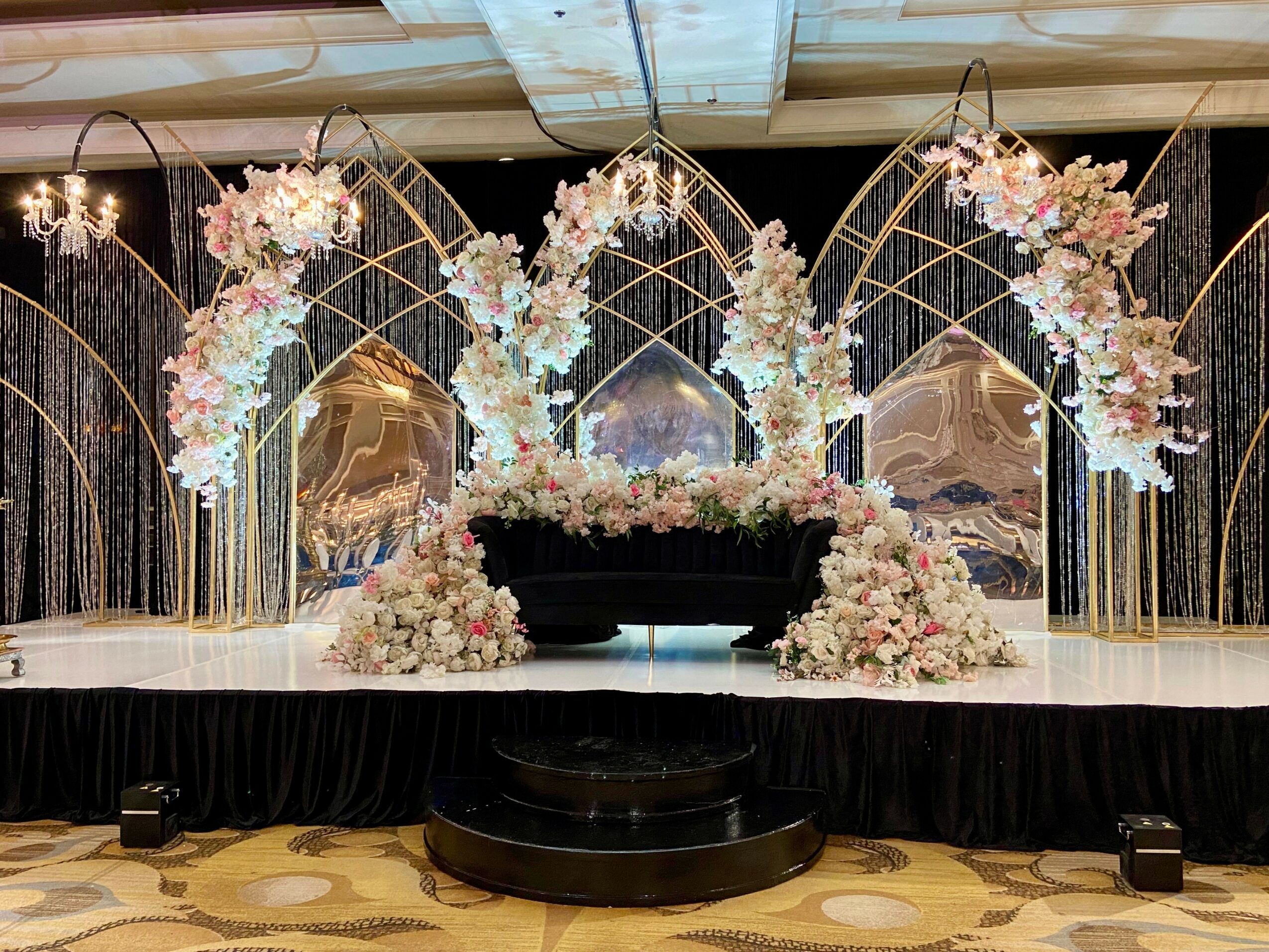 Decorative floral arrangement with gold arches and mirrors on a stage, black curtain backdrop, and floral clusters at base.