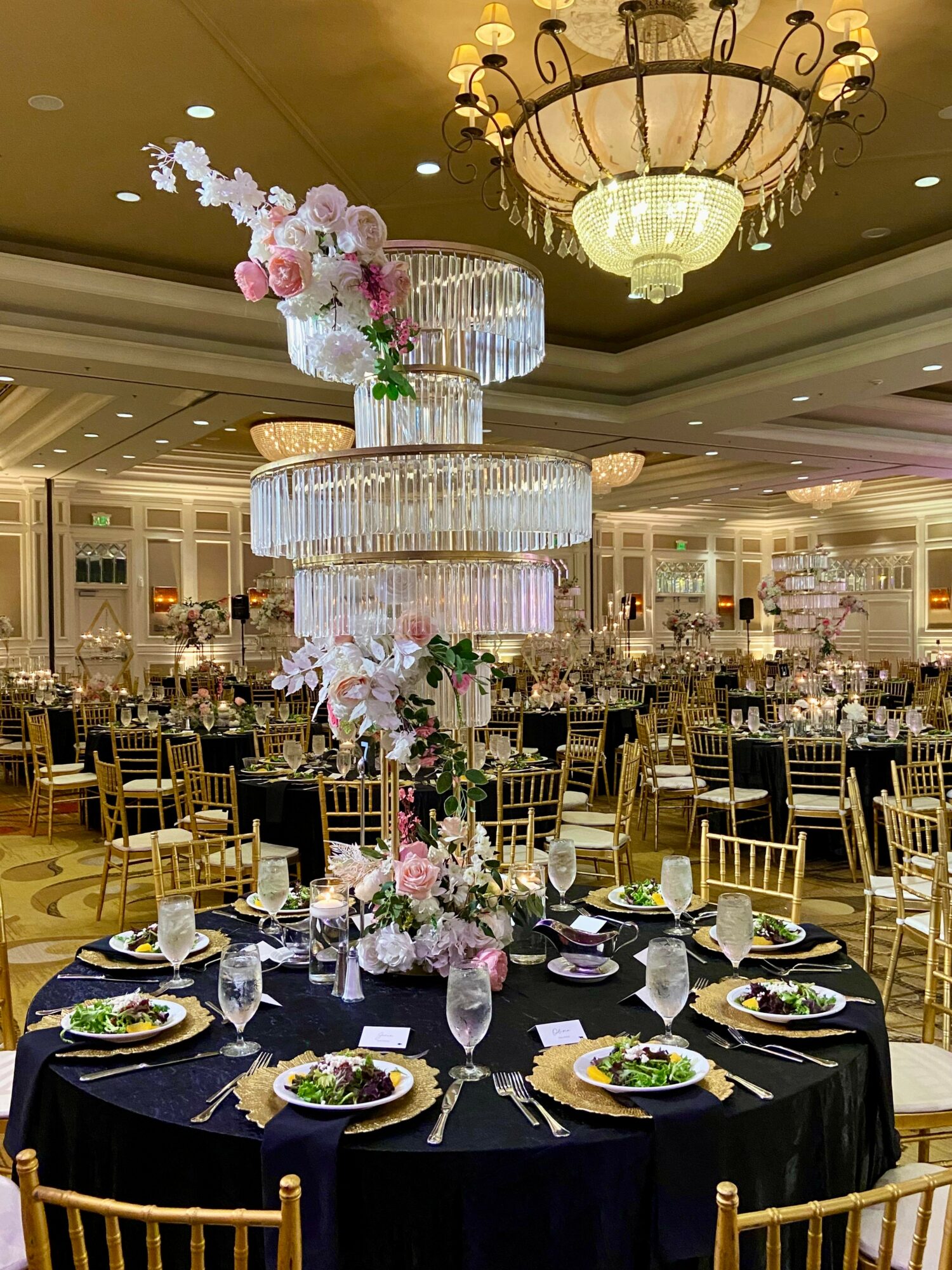 Elegant banquet table with a tall floral centerpiece and crystal chandelier in a decorated event hall.