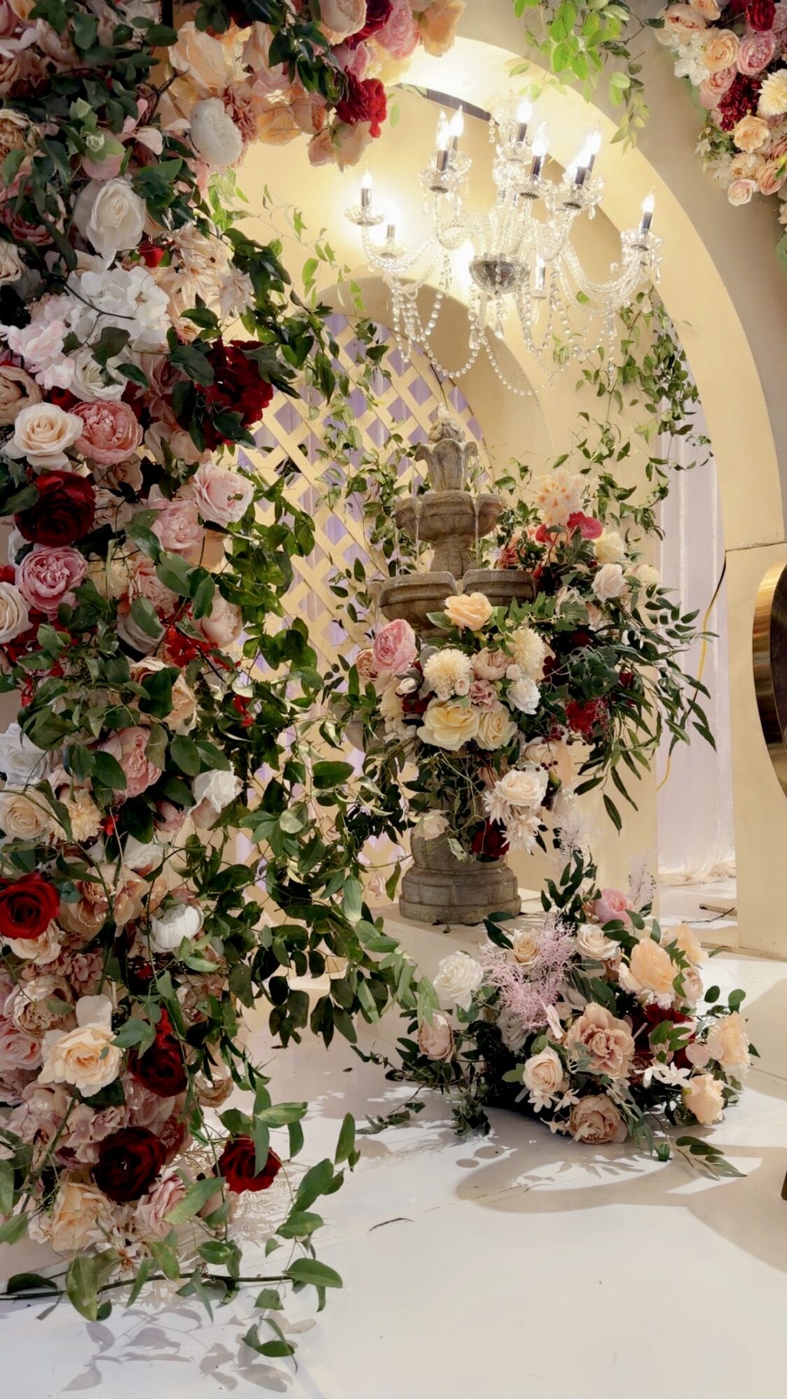 Decorative floral arch with pink, white, and red flowers, and a white backdrop with a chandelier, in an elegant setting.