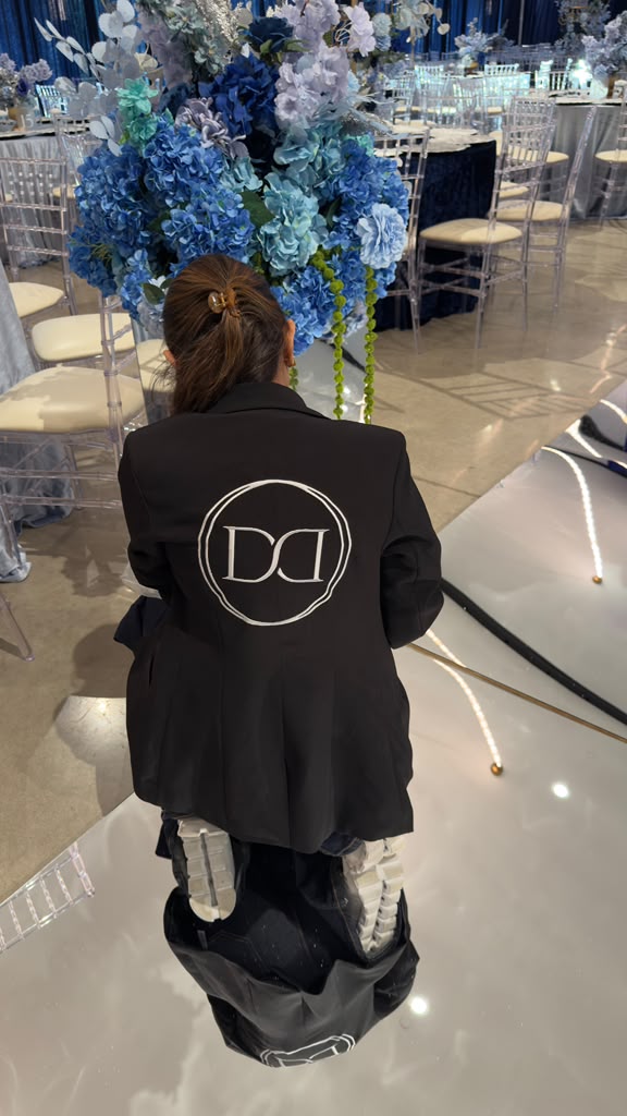 Person with brown hair in a bun, wearing a black jacket with a white logo on the back, kneeling in front of a floral arrangement.