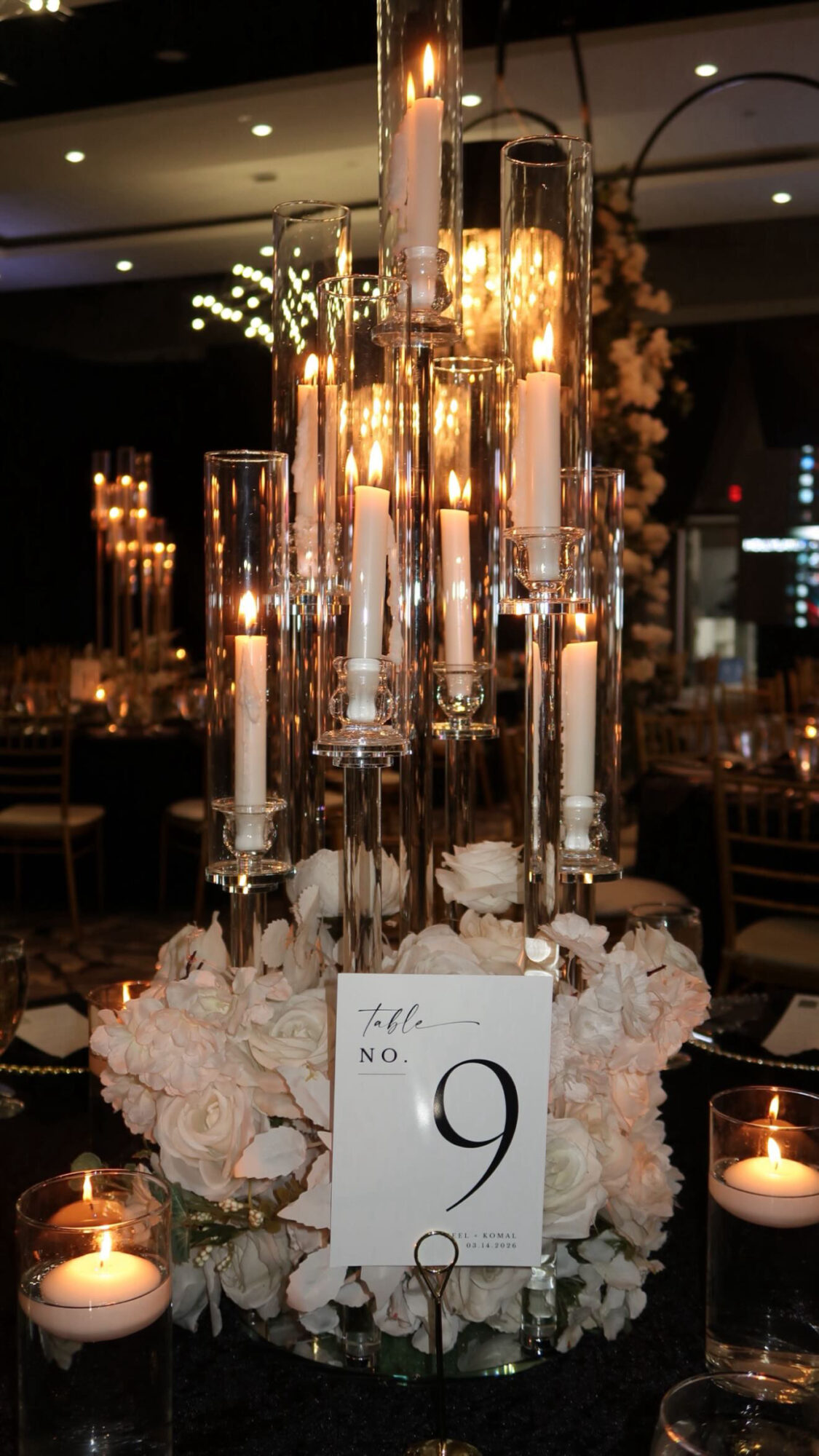 Elegant table centerpiece with tall candles, floral arrangement, and a table number card, surrounded by small lit candles.