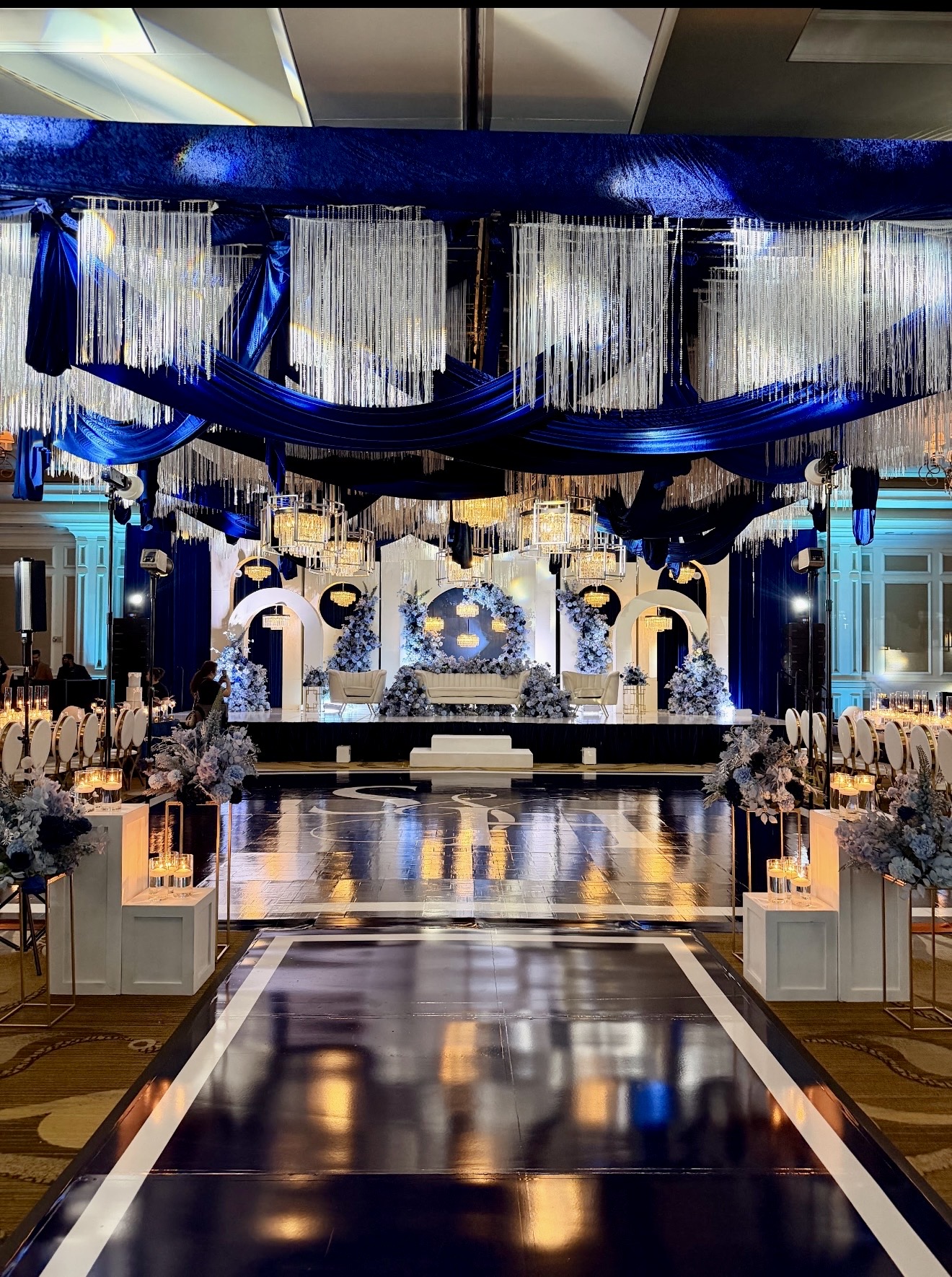 Elegant event stage with blue and white decorations, floral arrangements, and a reflective runway, in a grand hall.