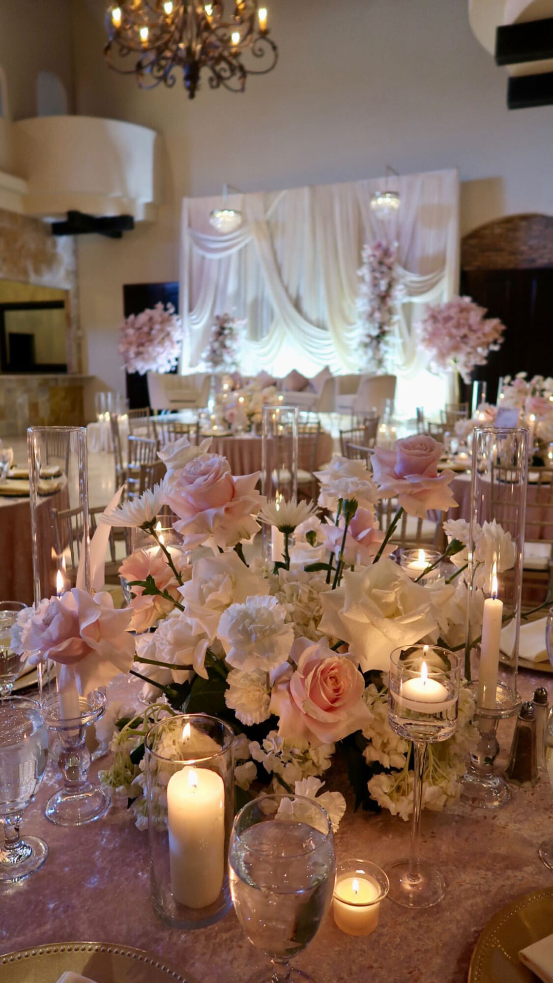 Elegant event table with pink and white flowers, candles, and a draped backdrop in a decorated room.