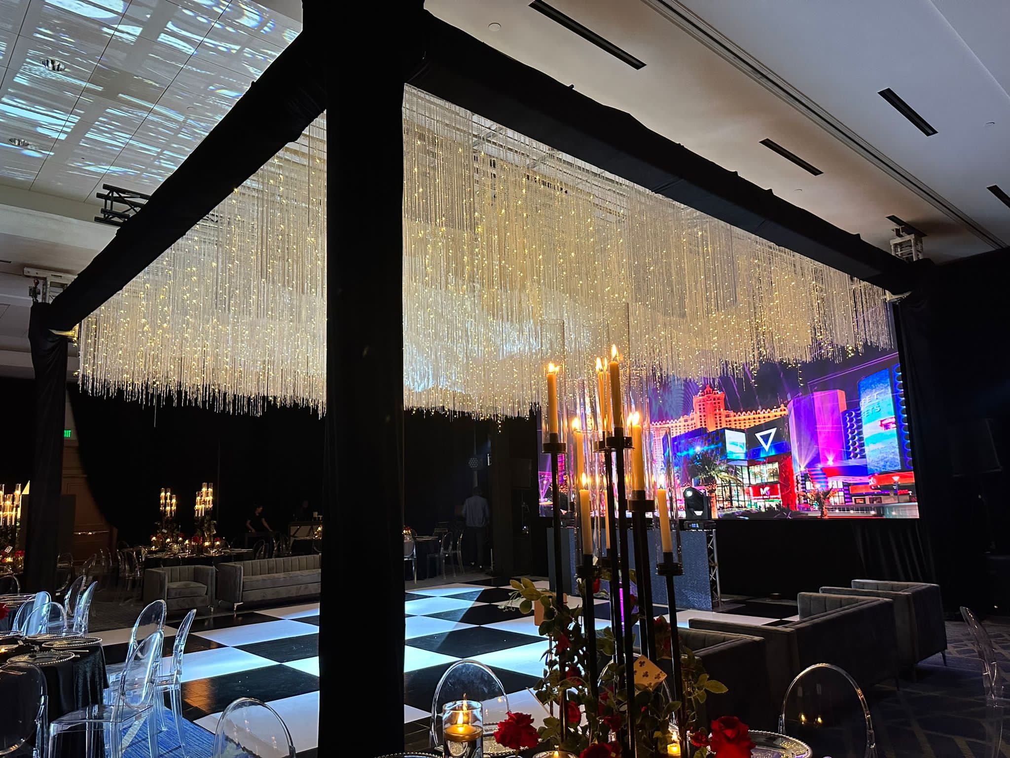 Indoor event space with a checkered dance floor, chandeliers, and city lights visible through large windows, nighttime scene.