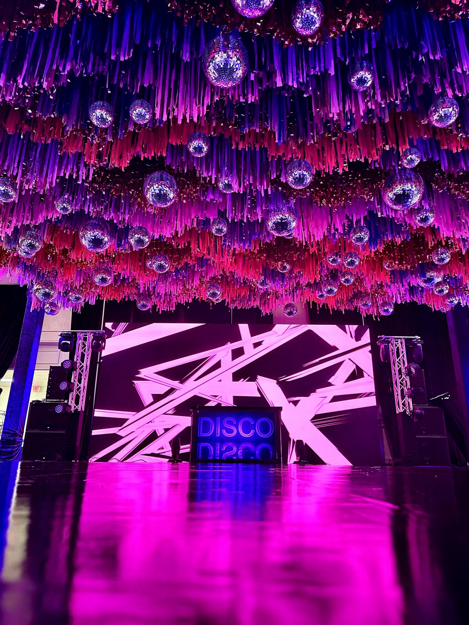 Stage with digital display and colorful hanging decorations, pink and purple lighting, and a shiny floor.