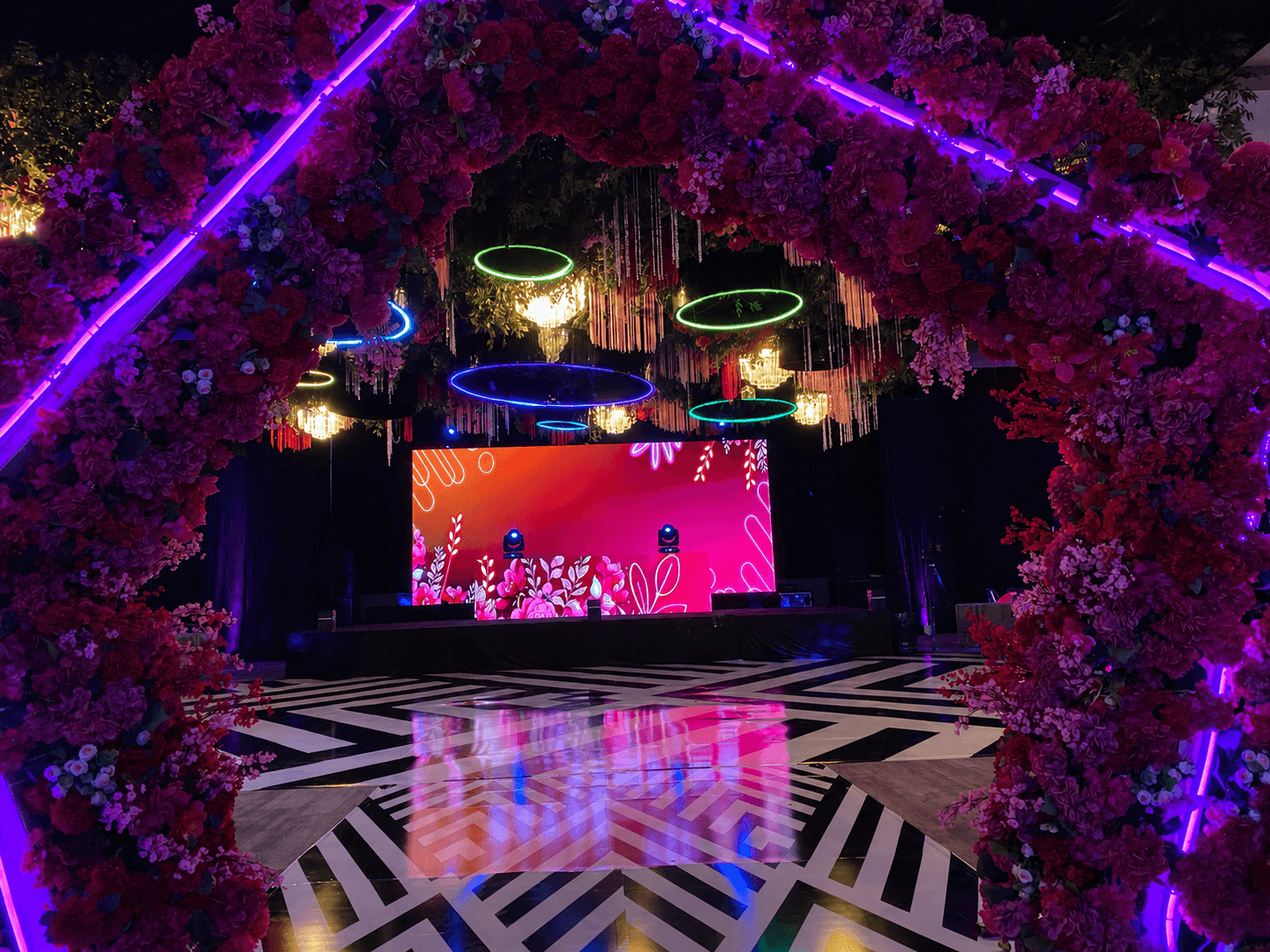 Stage with colorful background, surrounded by pink flowers and neon lights, reflecting on a patterned floor.