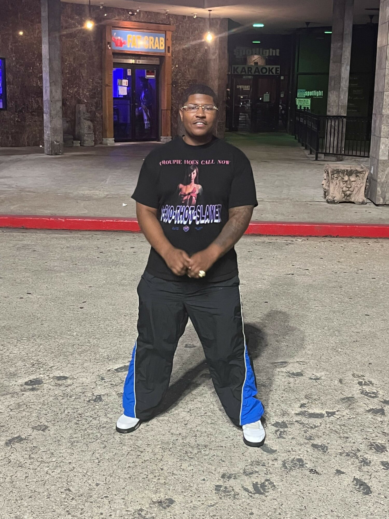 Young man standing on street at night, wearing black t-shirt, black pants with blue stripe, and white shoes.