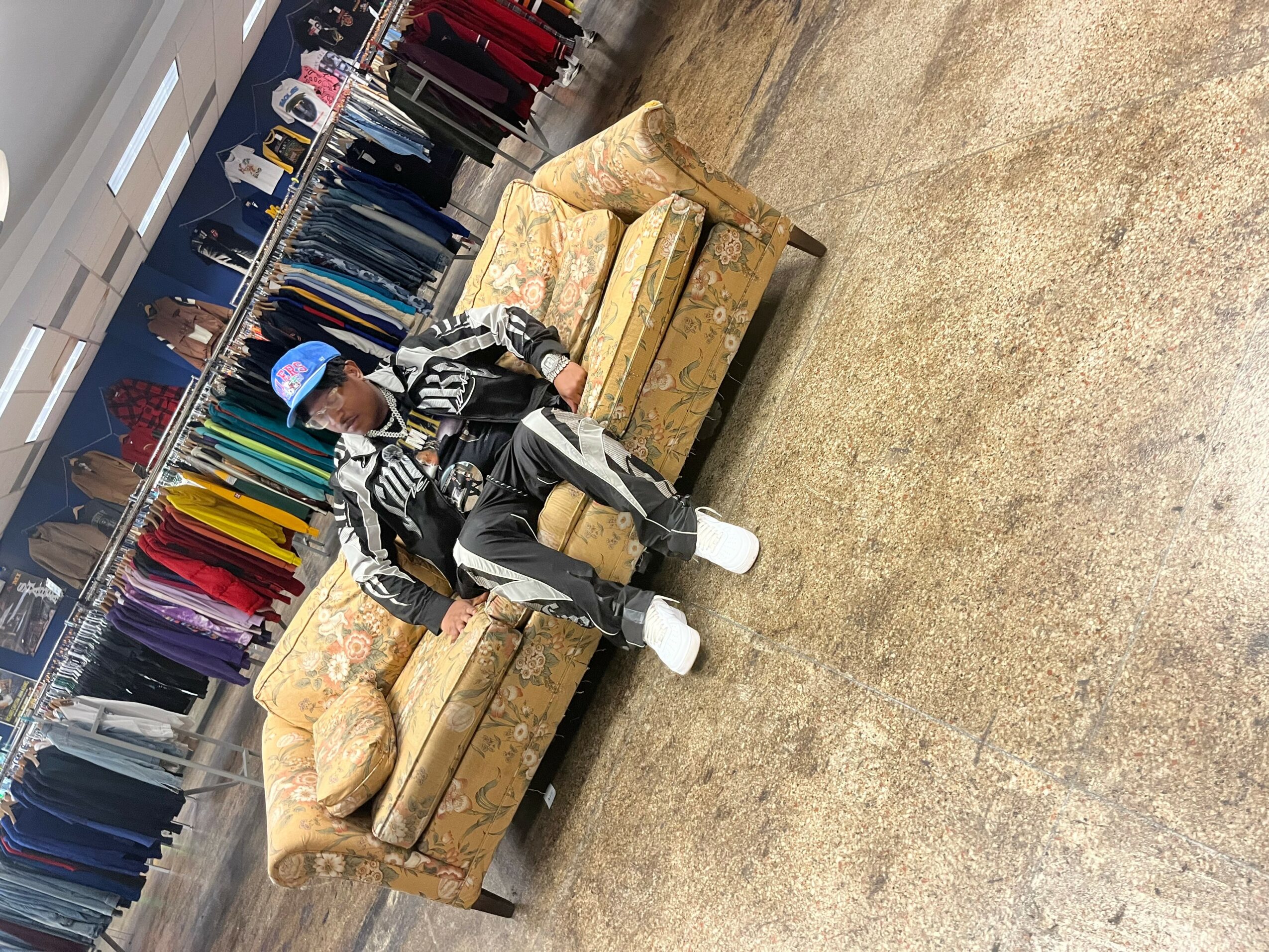 Person sitting on a sofa in a clothing store, surrounded by racks of colorful clothes, wearing a cap and casual clothing.