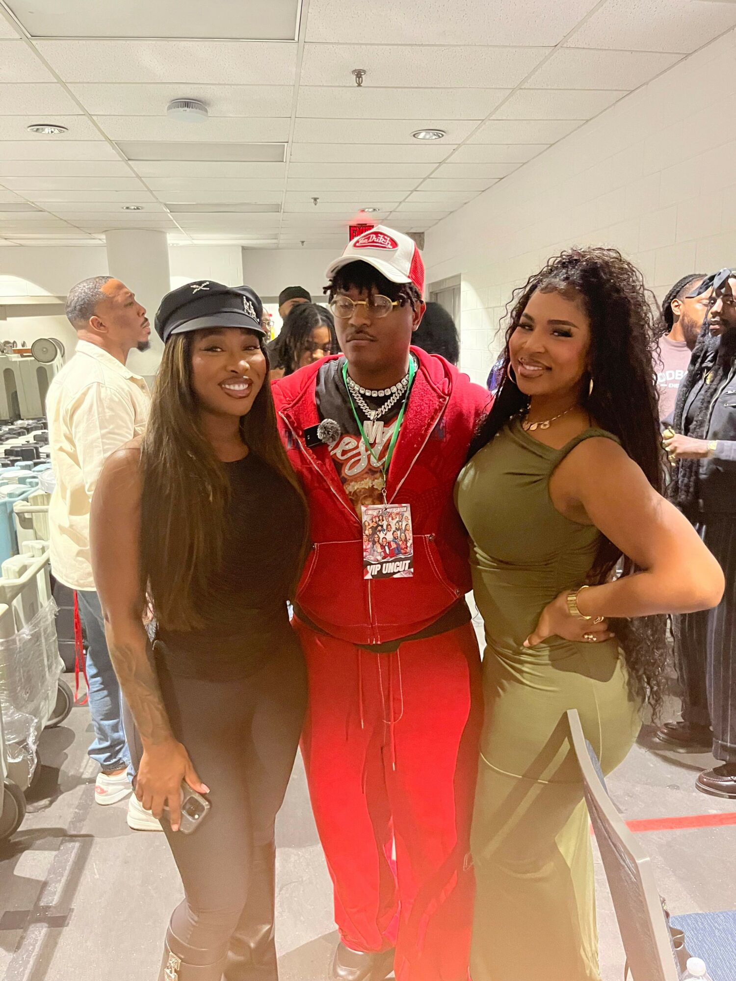 Three women standing together in a store, one in a red outfit, one in a green top, and one in a white shirt, with shelves in the background.
