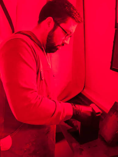 Person with glasses and beard using a laptop, in a red-lit room, facing right, with curtains and a screen nearby.