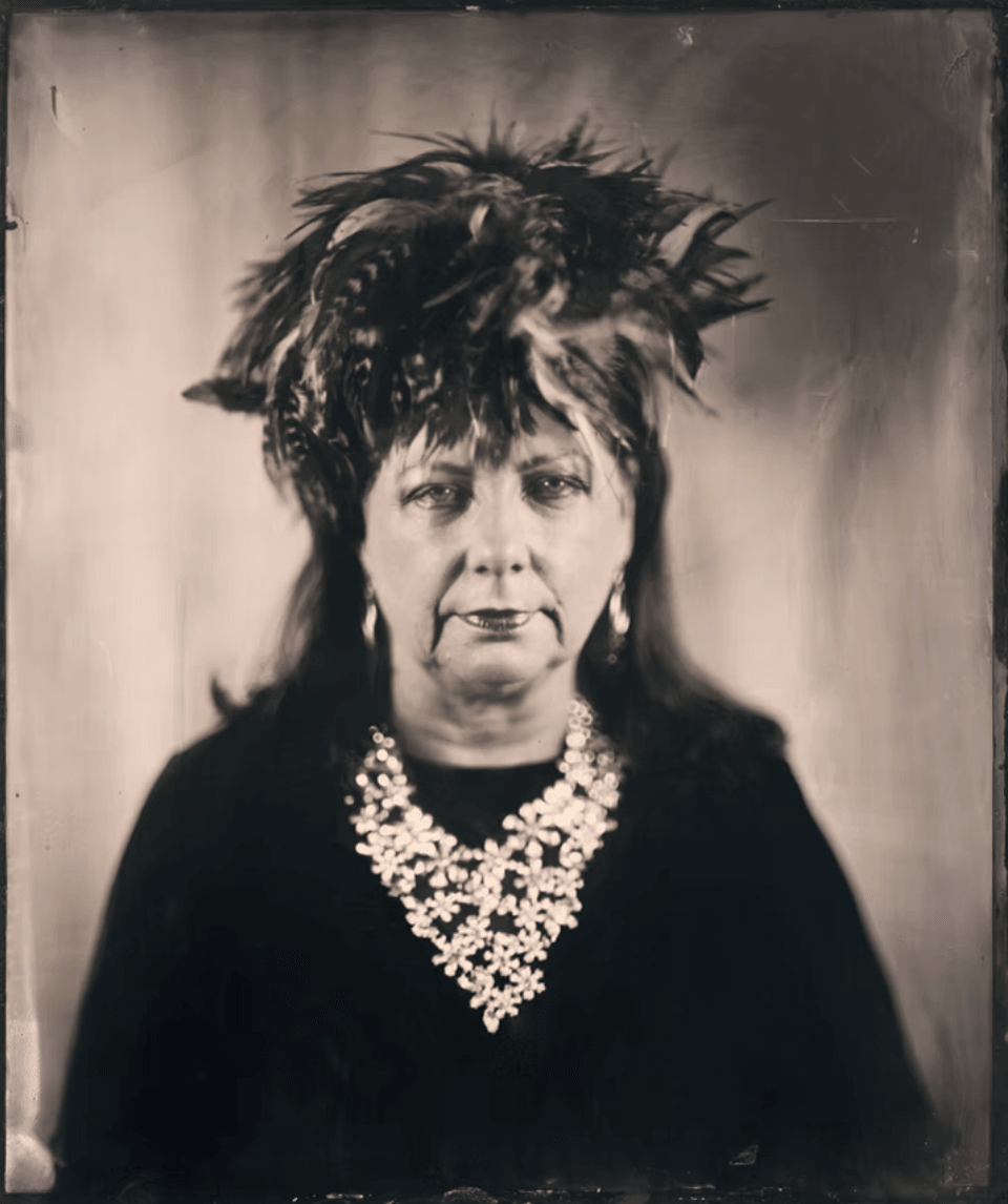 Black and white portrait of a woman with elaborate hairstyle and a large necklace, facing forward.