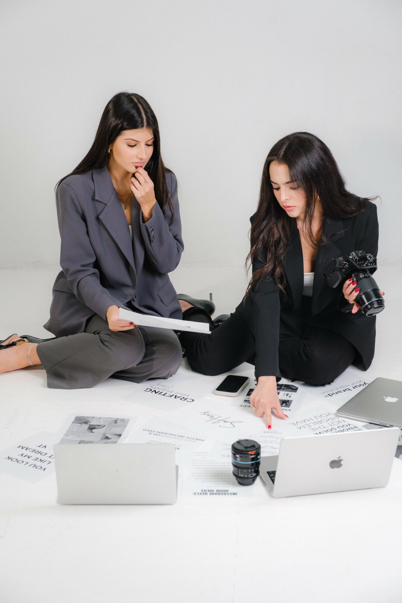 Two women sit on the floor with papers, laptops, and a camera, discussing and working together against a plain background.