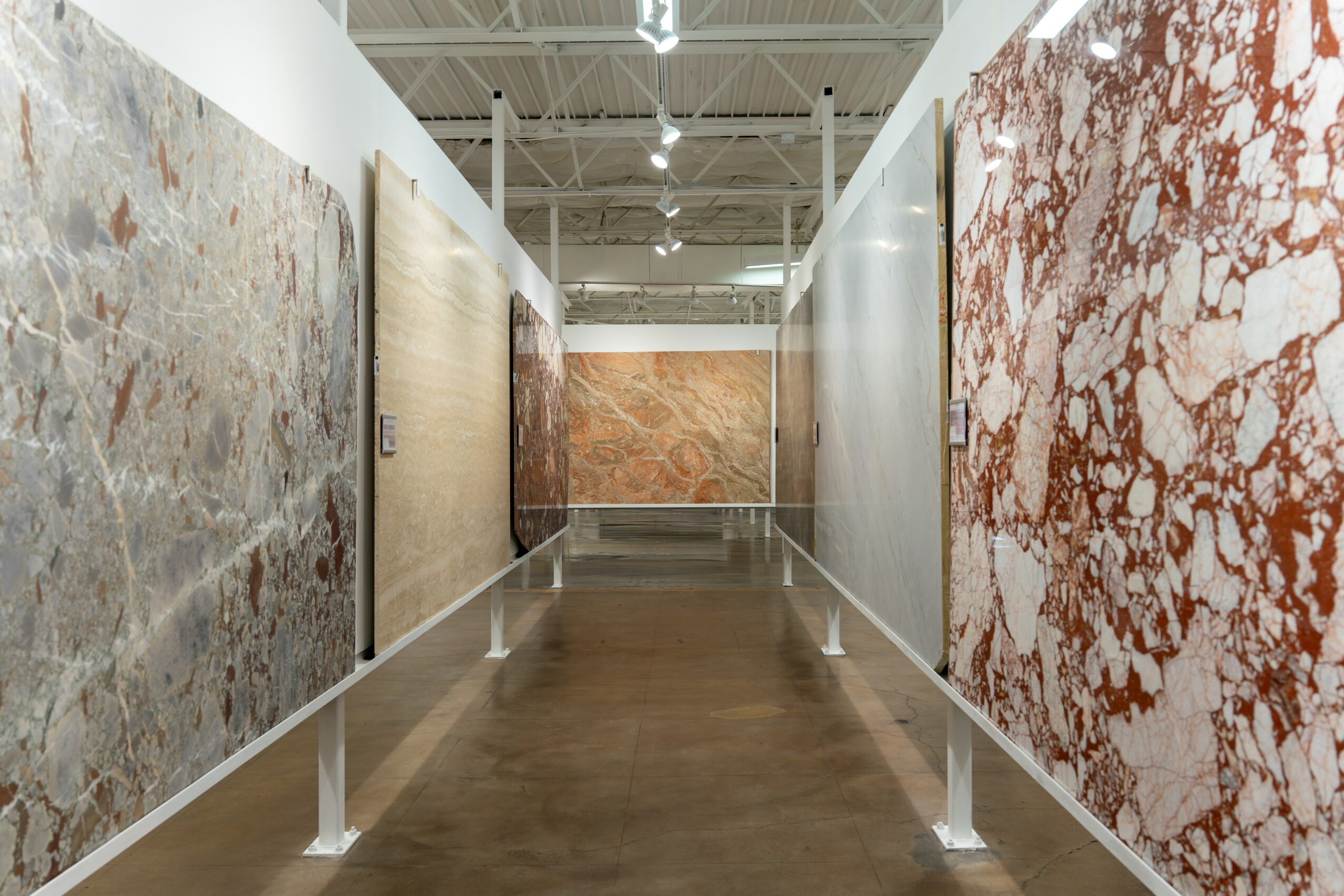 Gallery with large marble or stone slabs displayed on walls, wooden floor, high ceiling with lights, and white barriers.