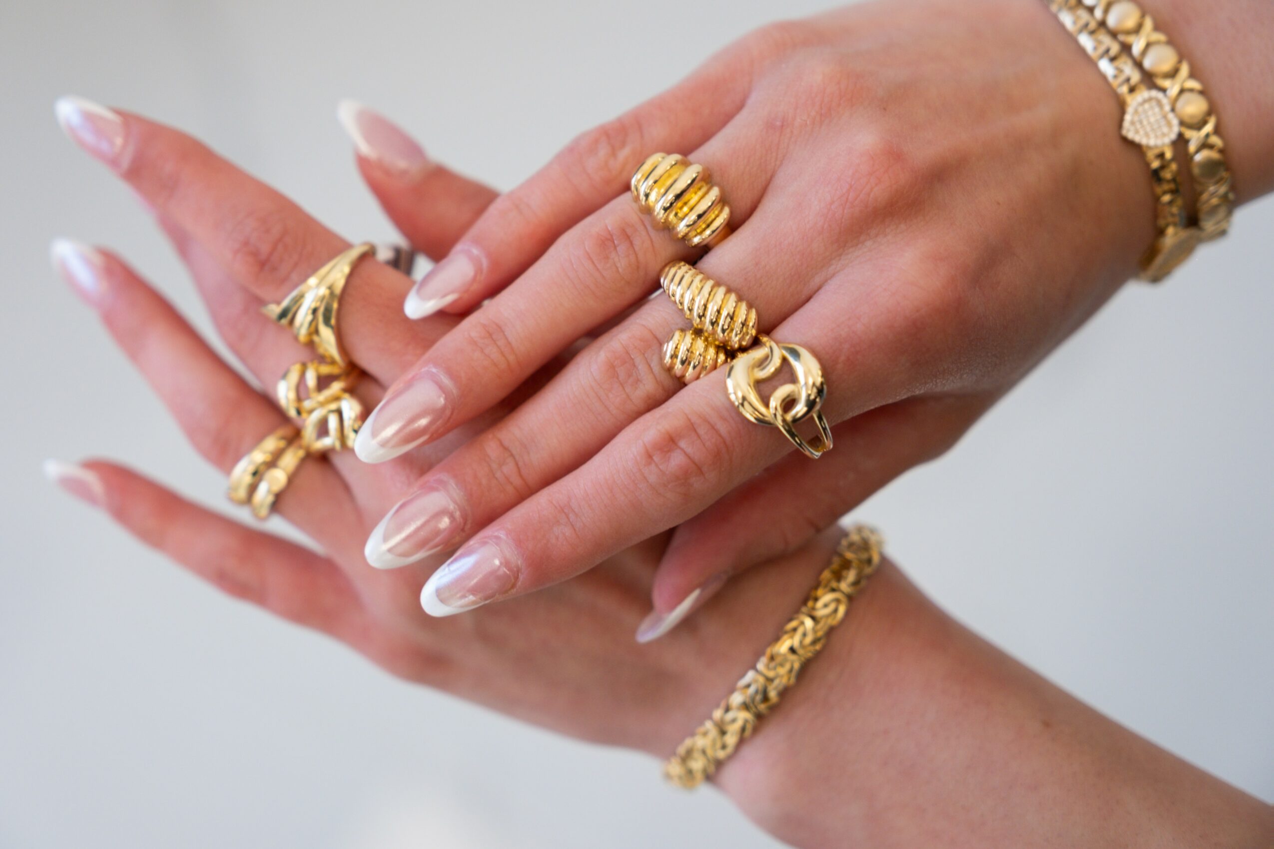 Hands with multiple gold rings and bracelets, with long, manicured nails, against a plain background.