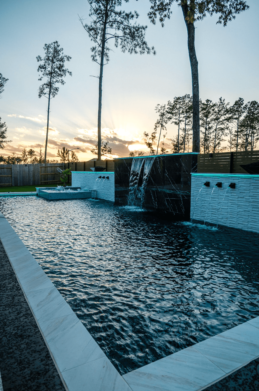 Swimming pool with waterfall feature, surrounded by trees and a fence, during sunset sky with clouds.