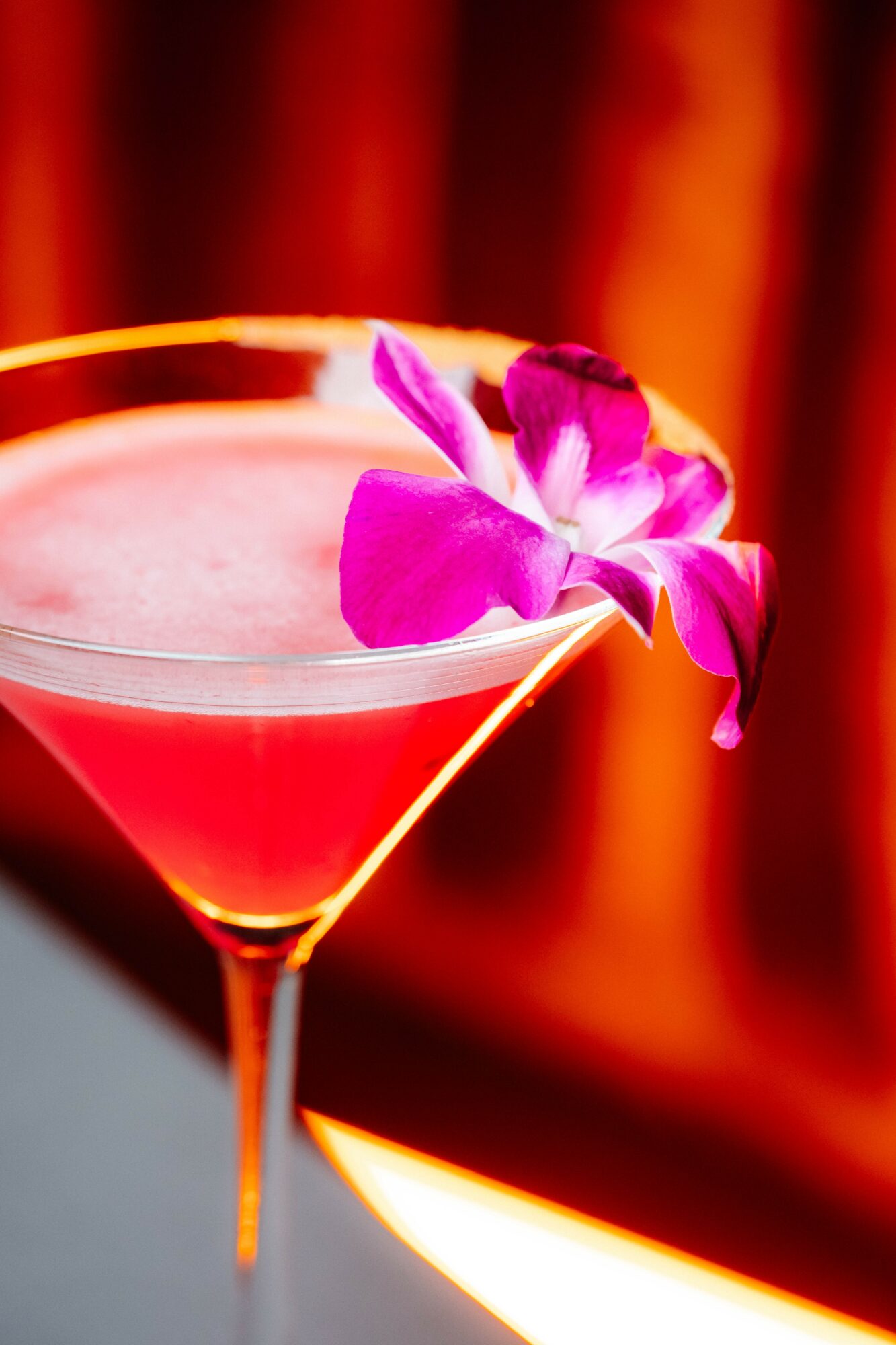 Pink cocktail in a martini glass with purple flowers garnish, against a warm, blurred background.