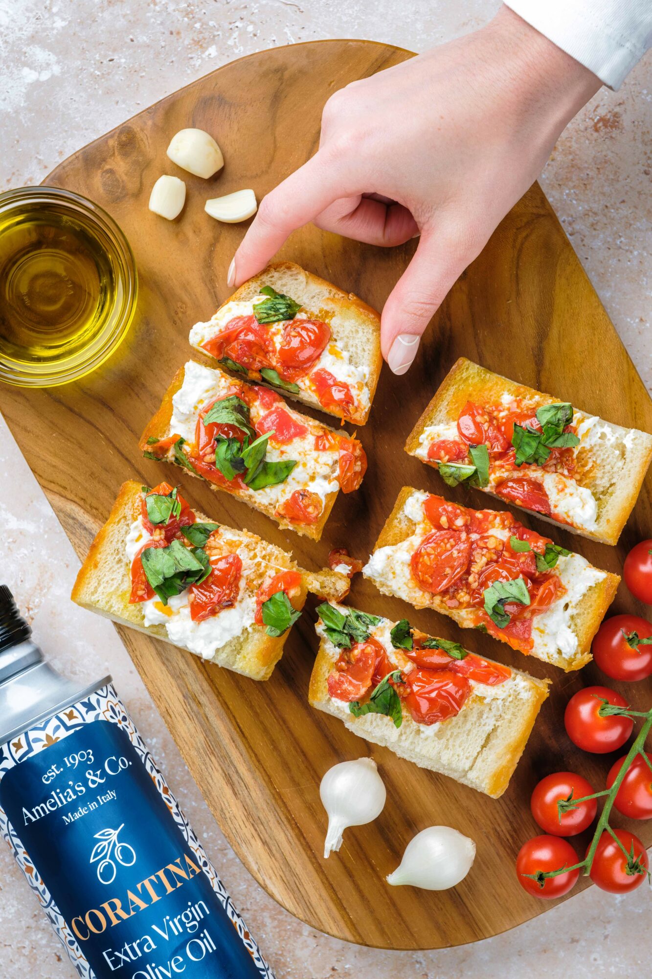 Hand reaching for tomato-topped bruschetta on a wooden platter, with garlic, olive oil, and cherry tomatoes nearby.