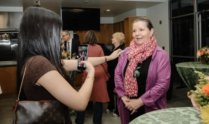 Two women talking at a social gathering, one smiling and wearing a pink scarf, the other holding a drink.