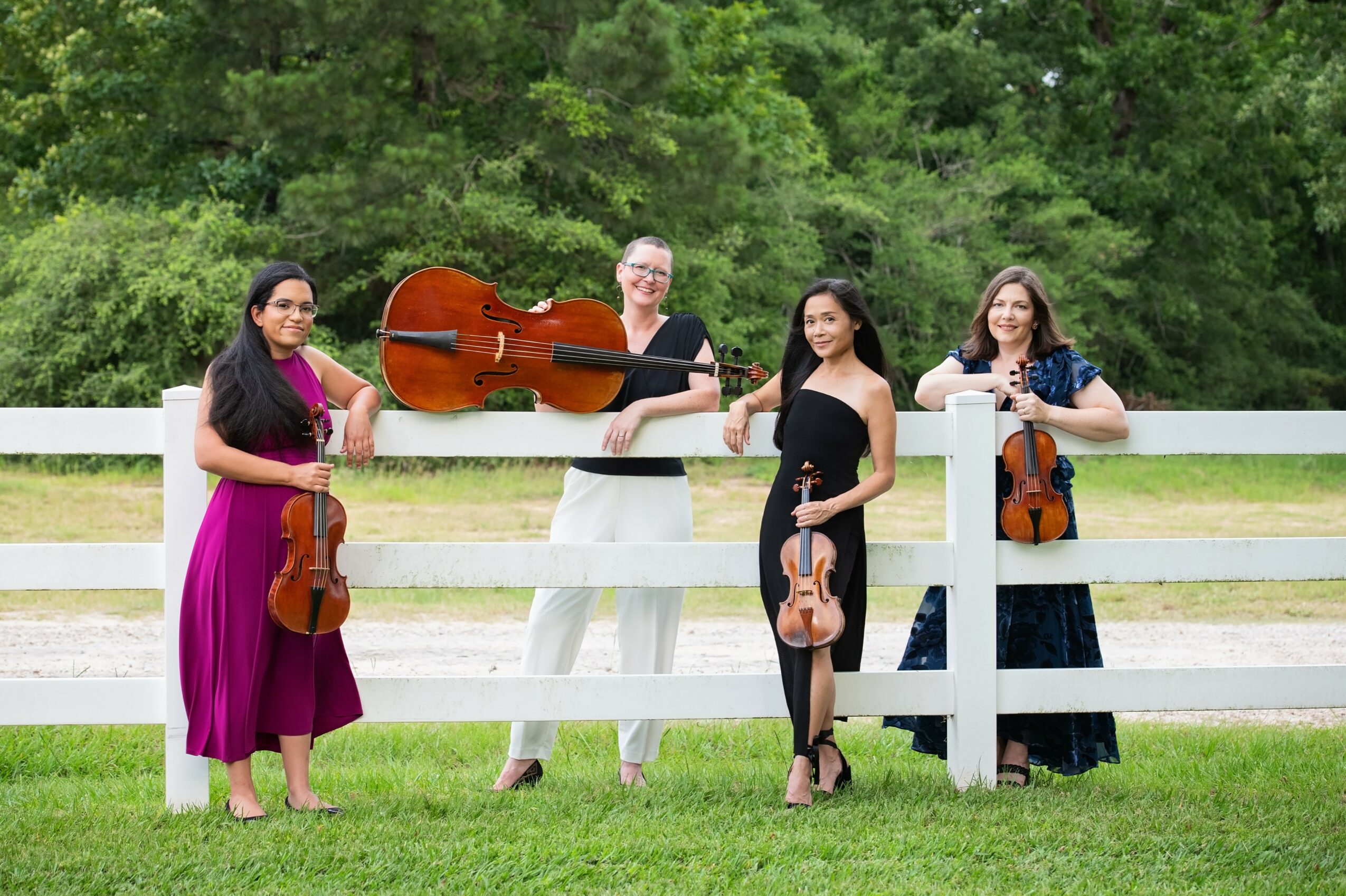 Four women with musical instruments standing outdoors on grass, with trees and a white fence in the background.