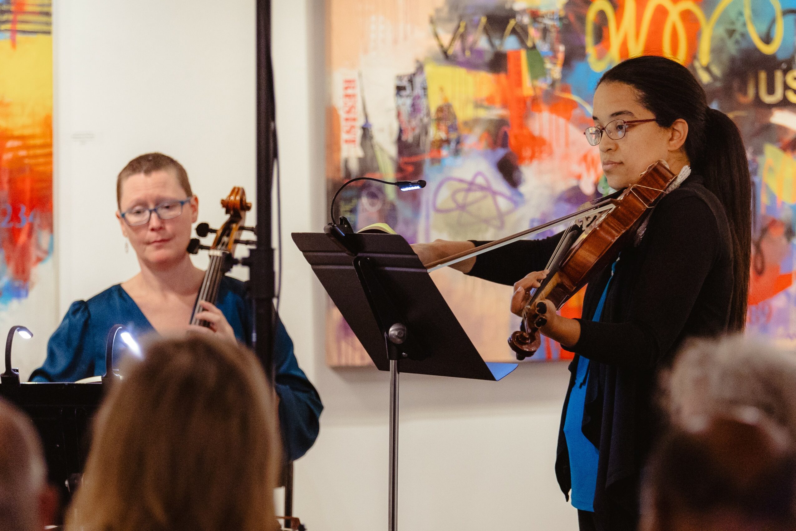 Two women playing violins in an art gallery, with an audience watching, colorful artwork on the wall behind them.